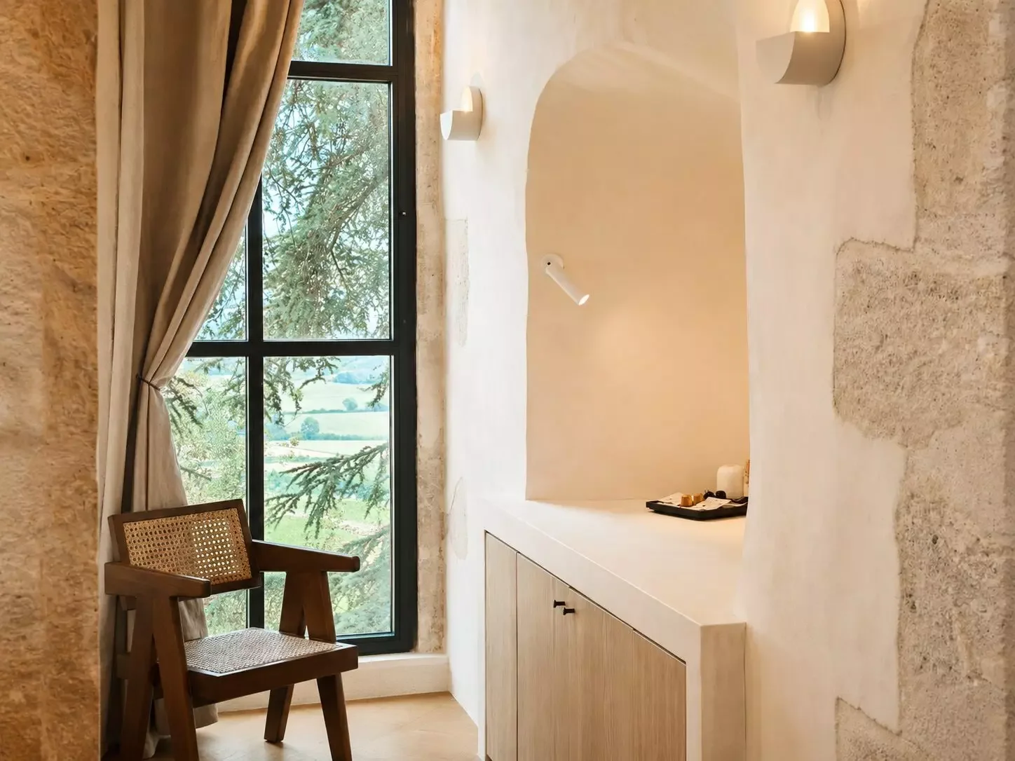 Une pièce lumineuse avec un grand fenêtre, un fauteuil en bois avec un siège en canevas, et un meuble en bois avec une surface blanche. La fenêtre laisse entrer la lumière naturelle et offre une vue sur des arbres et des champs. La pièce a des murs e