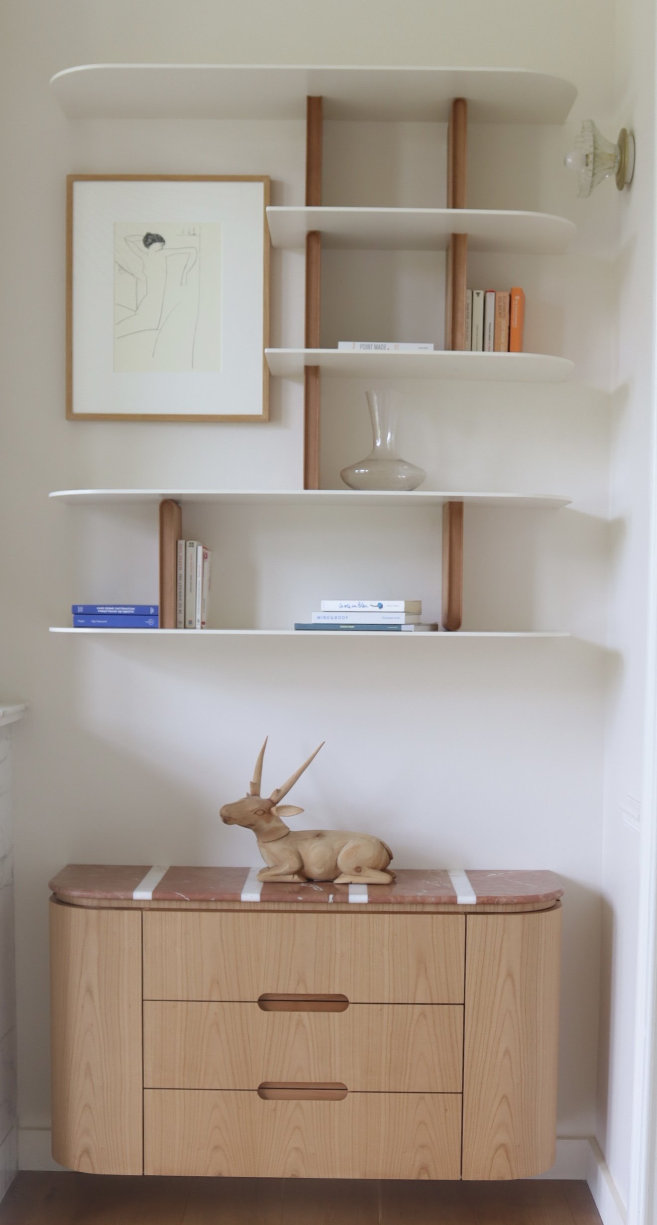 Une étagère blanche à rayures en bois avec des livres, une oeuvre d'art et une lampe, au-dessus d'une commode en bois clair avec une sculpture de renard en bois.