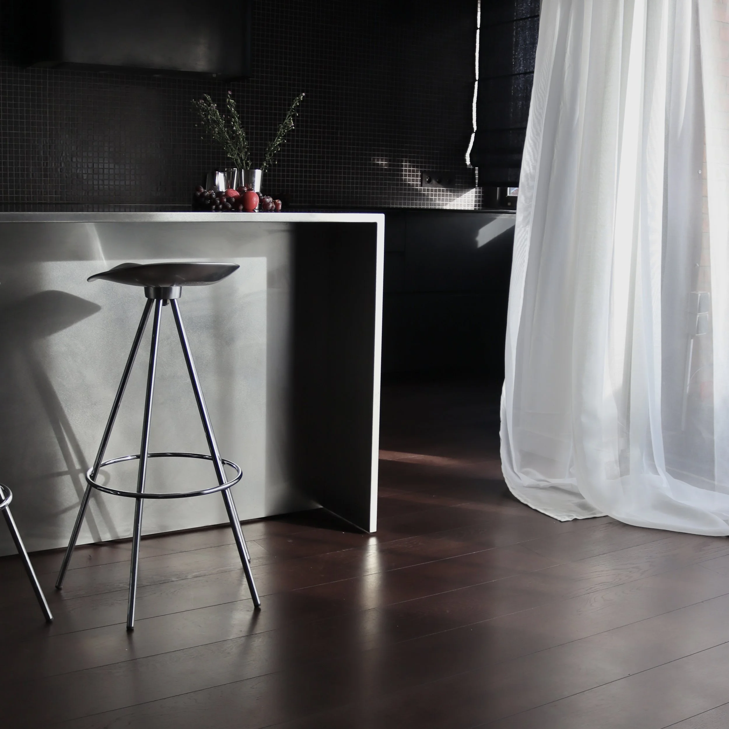 Une cuisine moderne avec un plan de travail en métal, une chaise haute en métal, un mur noir avec de petites mosaïques, des vases avec des fleurs et des fruits rouges, une fenêtre avec des rideaux blancs légers.