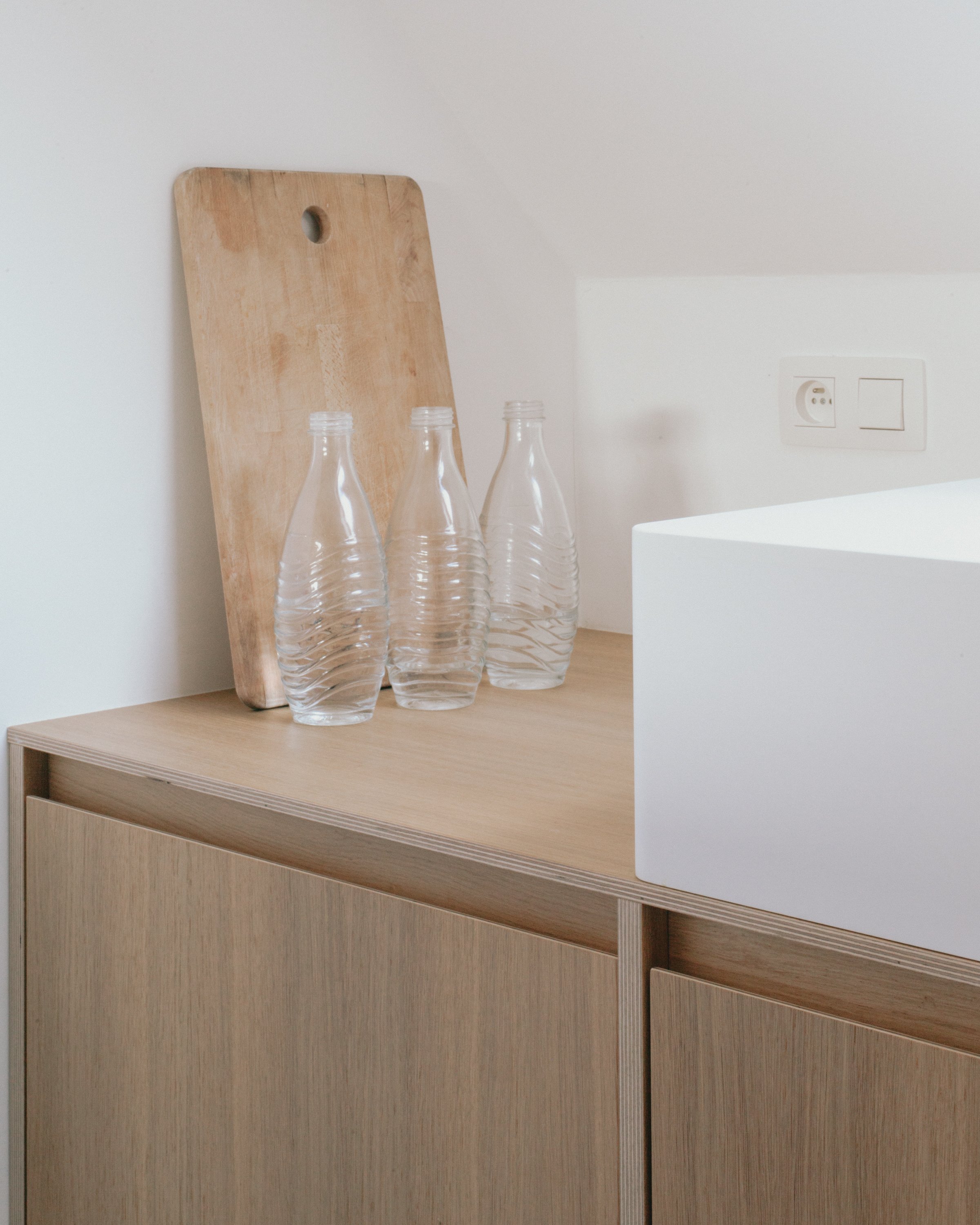 Une planche en bois posée contre un mur blanc, avec trois bouteilles en plastique vides sur une surface en bois clair. À droite, une prise électrique encastrée dans le mur et une surface blanche.