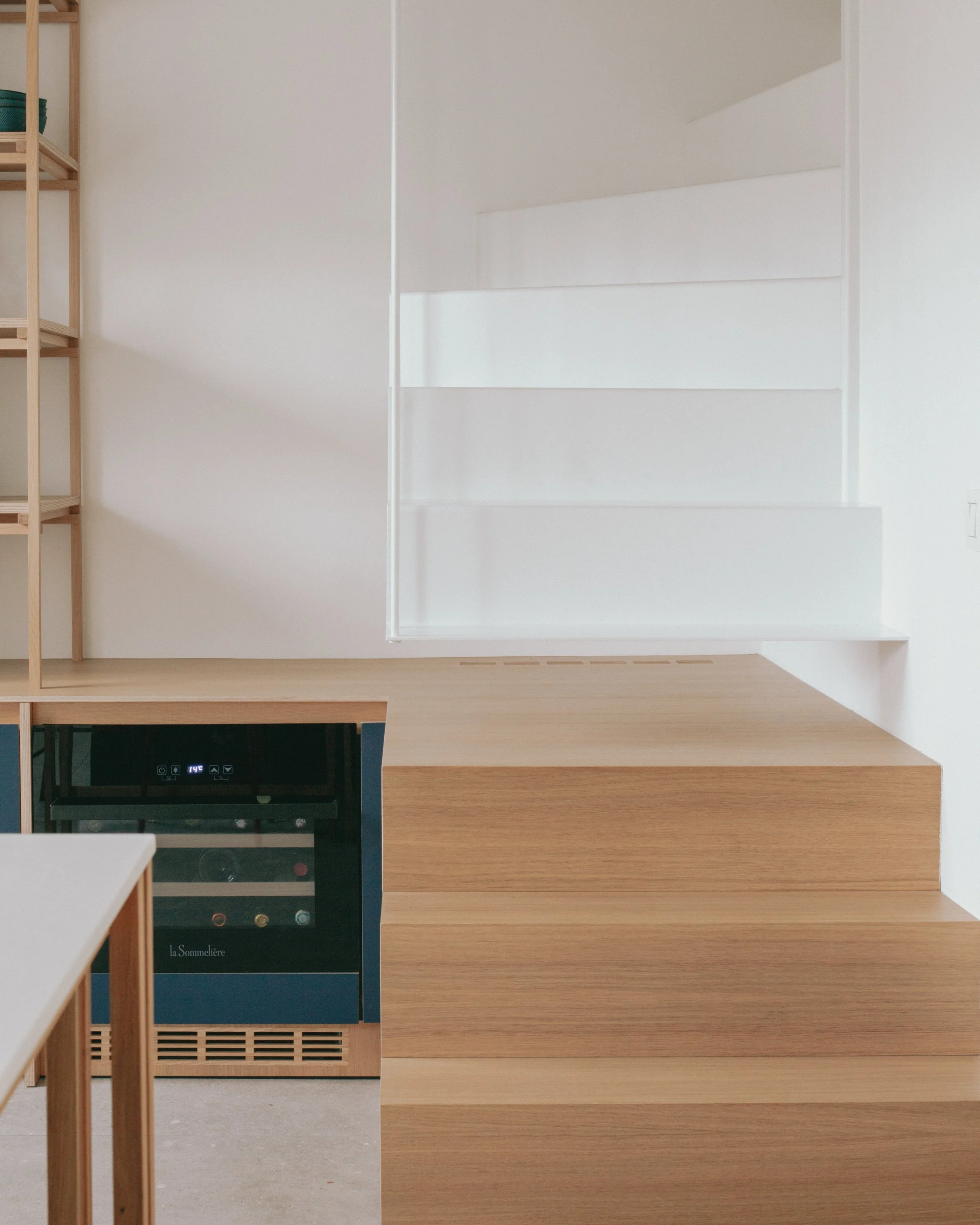 Intérieur moderne avec étagères en bois clair, un meuble en bois clair, et un électroménager noir avec un écran digital. Murs blancs et sol en béton léger.
