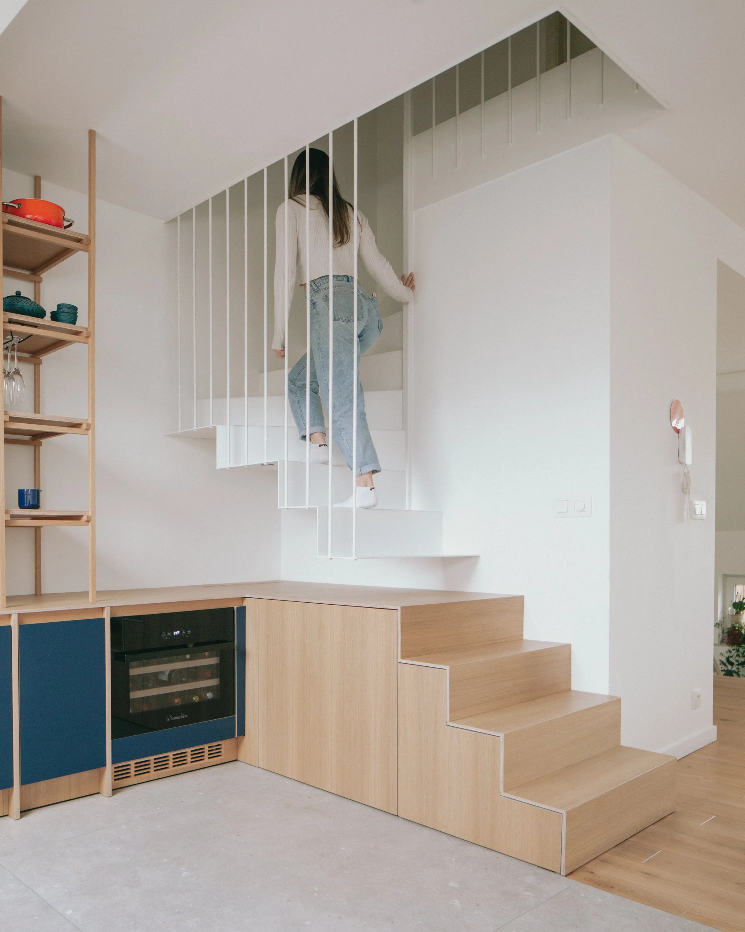 Une femme monte un escalier ludique en bois avec des marches en forme de blocs de différentes tailles, intégrée dans un espace intérieur moderne avec des murs blancs et une unité de rangement en bois.