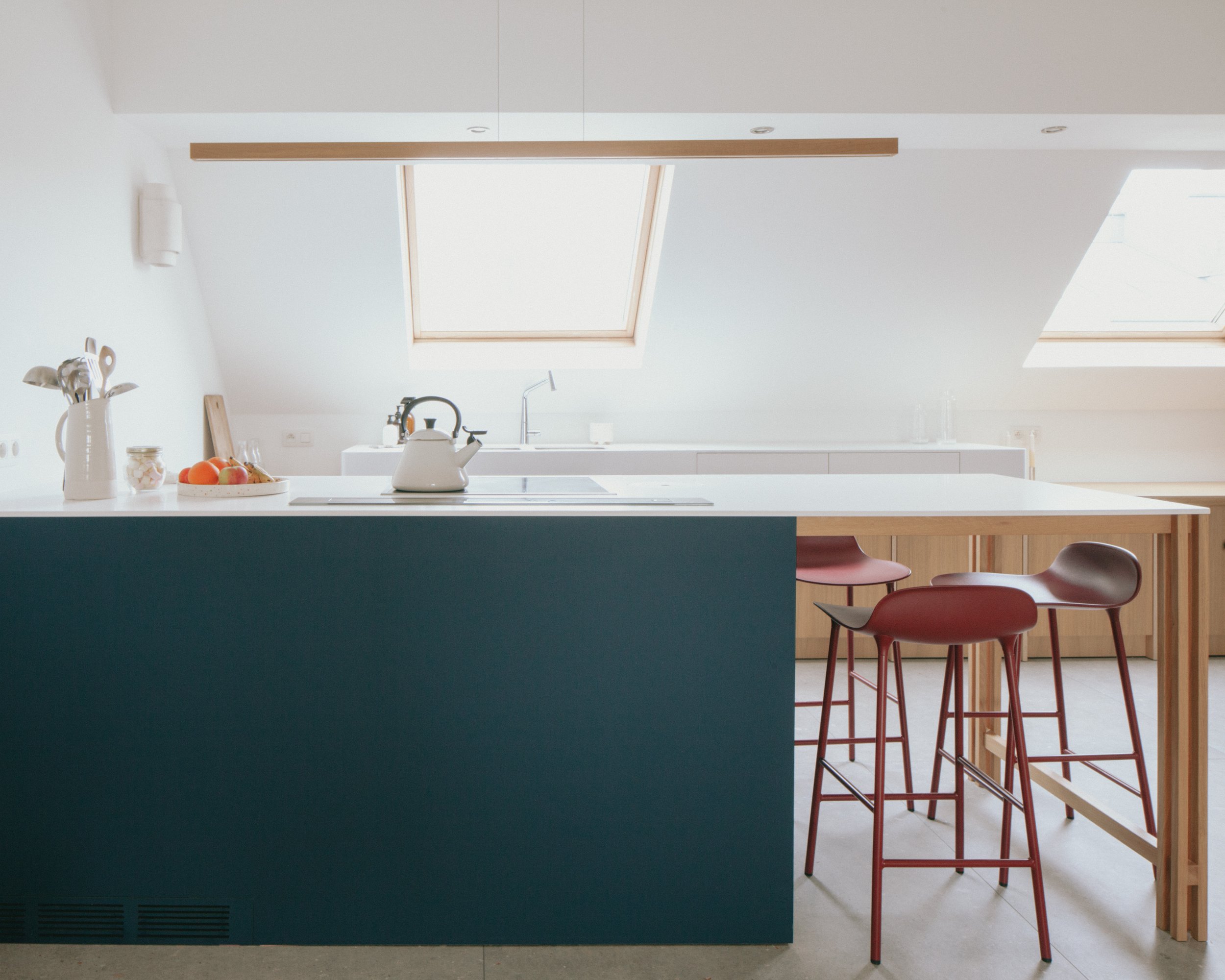 Cuisine moderne avec éclairage naturel venant d'une fenêtre de plafond, îlot central avec une théière blanche, un bol de pommes et des ustensiles dans un vase, chaises rouges autour d'une table en bois.