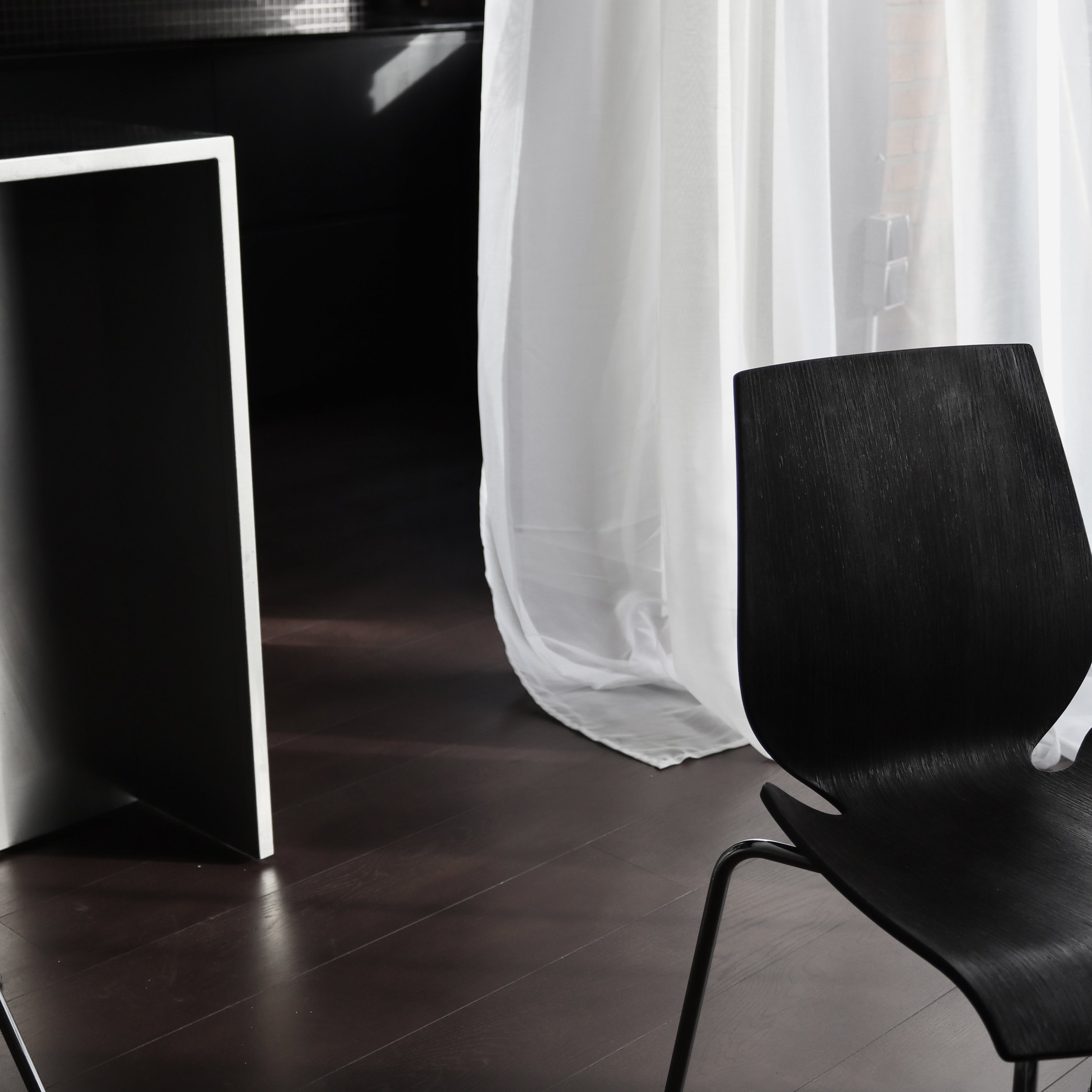 Une pièce avec un sol en bois foncé, une fenêtre avec des rideaux blancs semi-transparents, une chaise noire en plastique ou en cuir et une étagère noire.