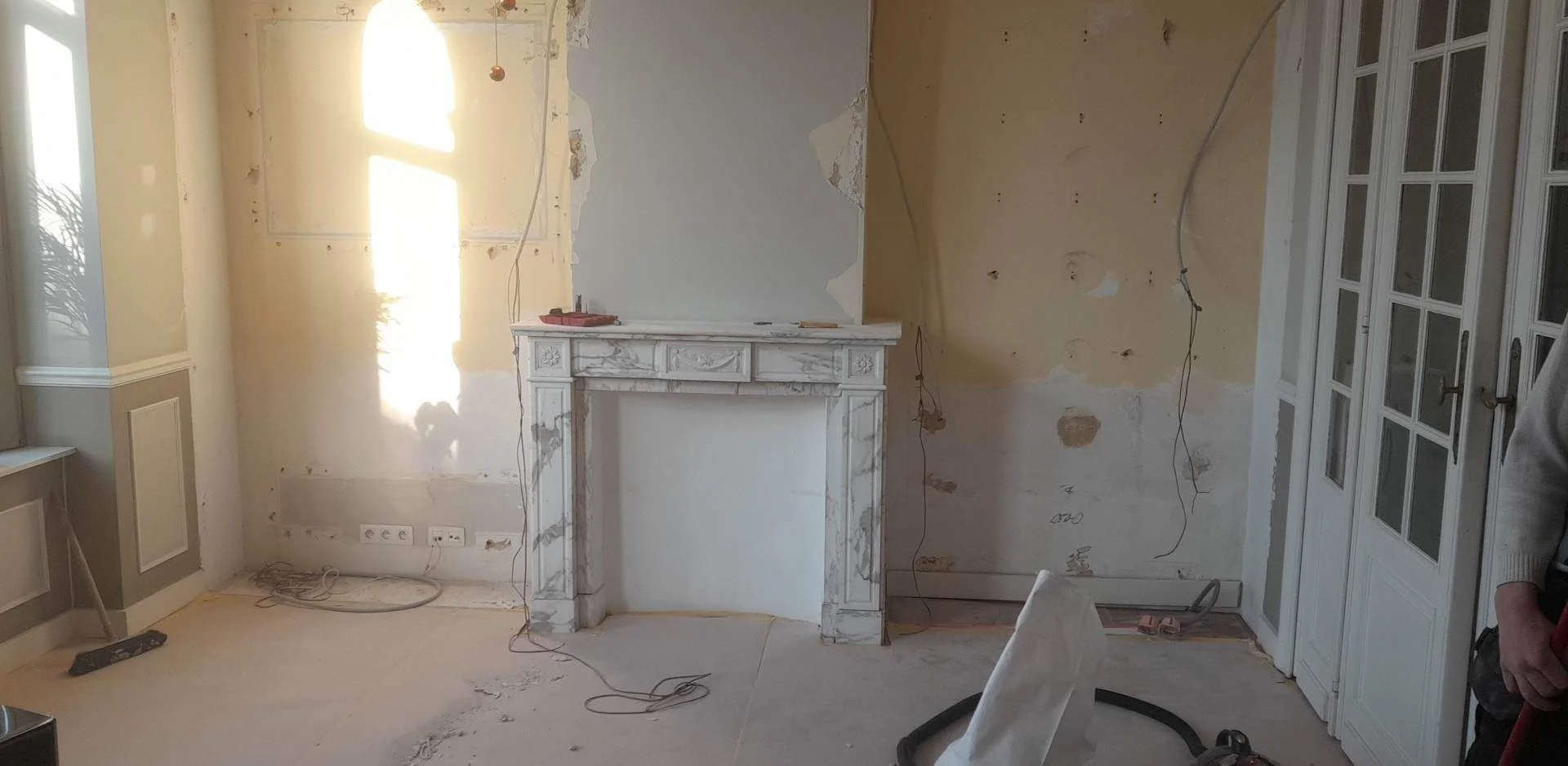 Une pièce en cours de rénovation avec un mur en train d'être retapissé, une cheminée en marbre, des câbles électriques suspendus, et des meubles en cours de montage ou de dépose.