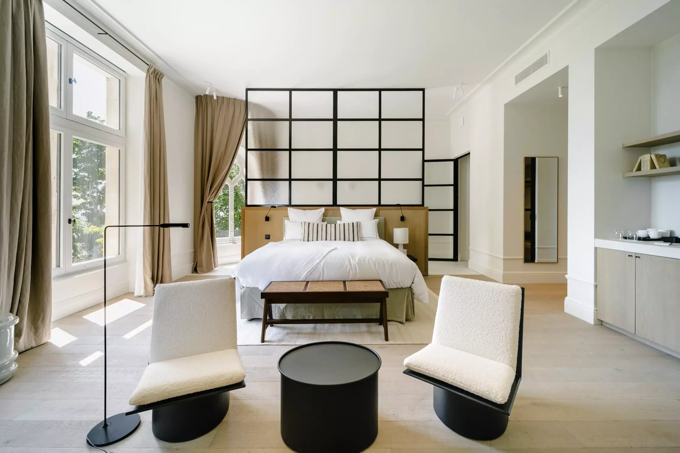 Chambre lumineuse avec lit, fenêtres, rideaux beige, division en verre noir, meubles modernes, chaise blanche, table ronde noire.