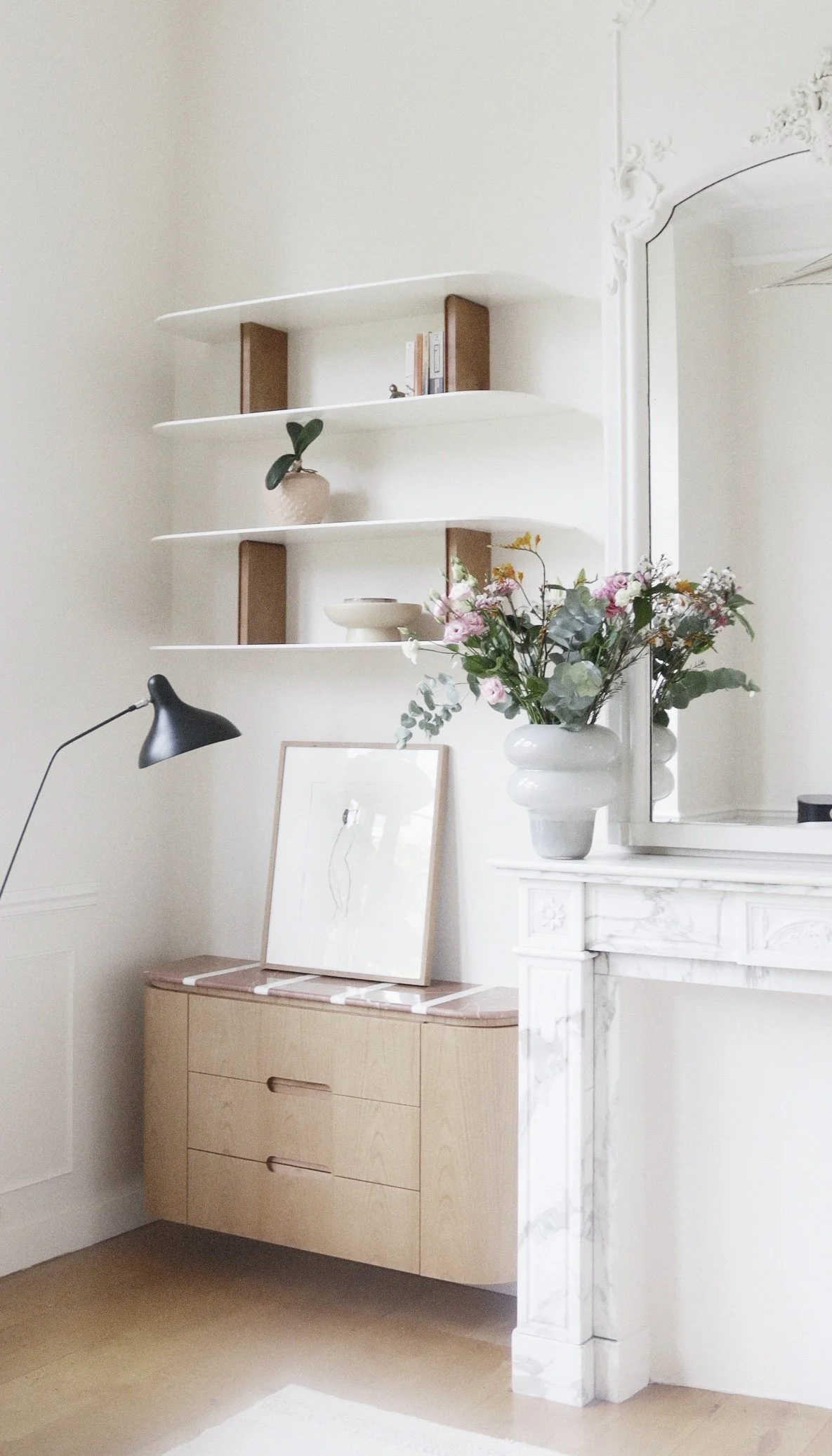 Intérieur minimaliste avec un meuble en bois clair, une lampe noire arquée, un cadre photo au sol, des étagères flottantes avec quelques livres et une plante, une grande vase avec des fleurs sur une cheminée en marbre blanc et un grand miroir.