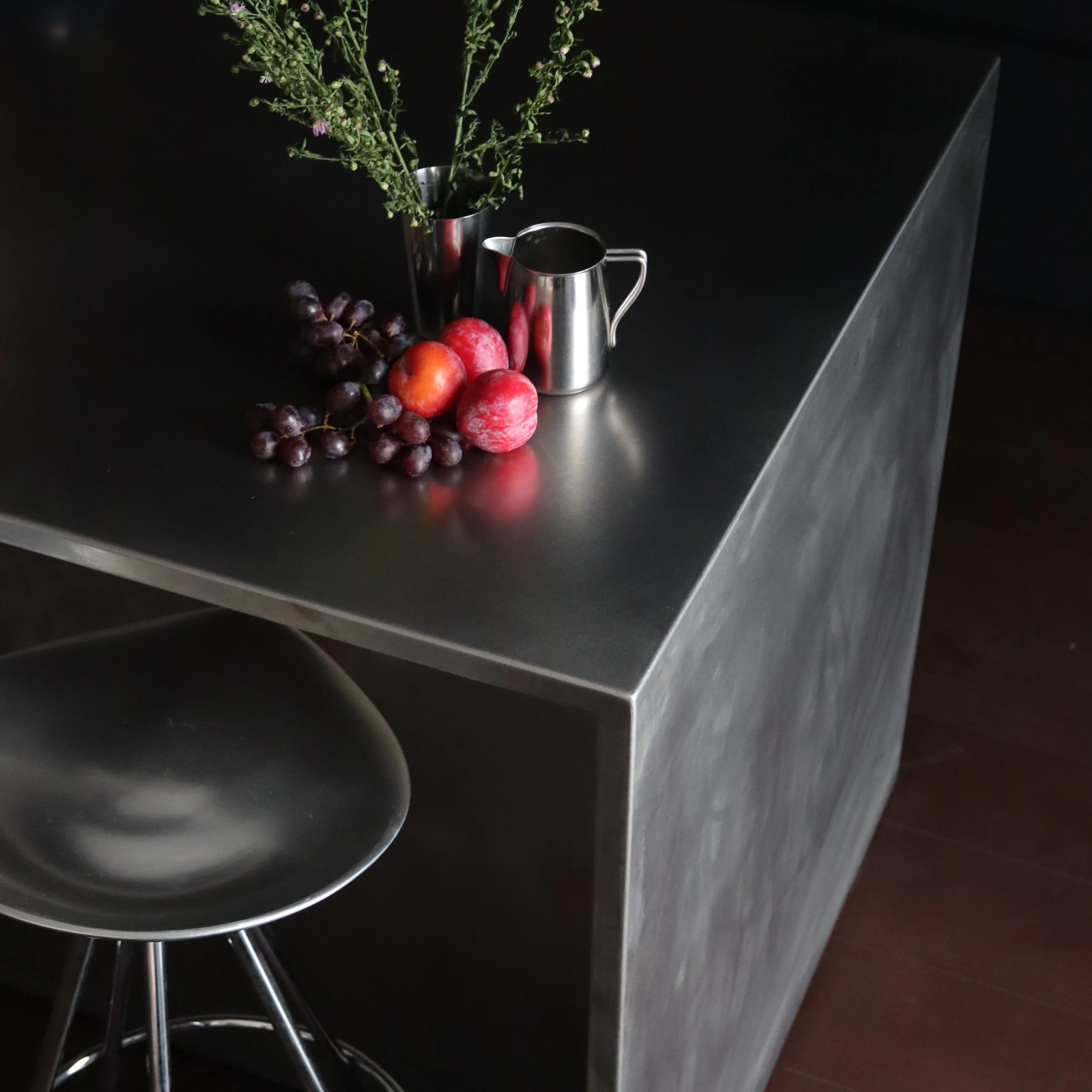 Une composition artistique avec des fruits tels que des raisins, des pommes, un poire et des plantes vertes dans deux vases en métal, placée sur une surface en métal noir.