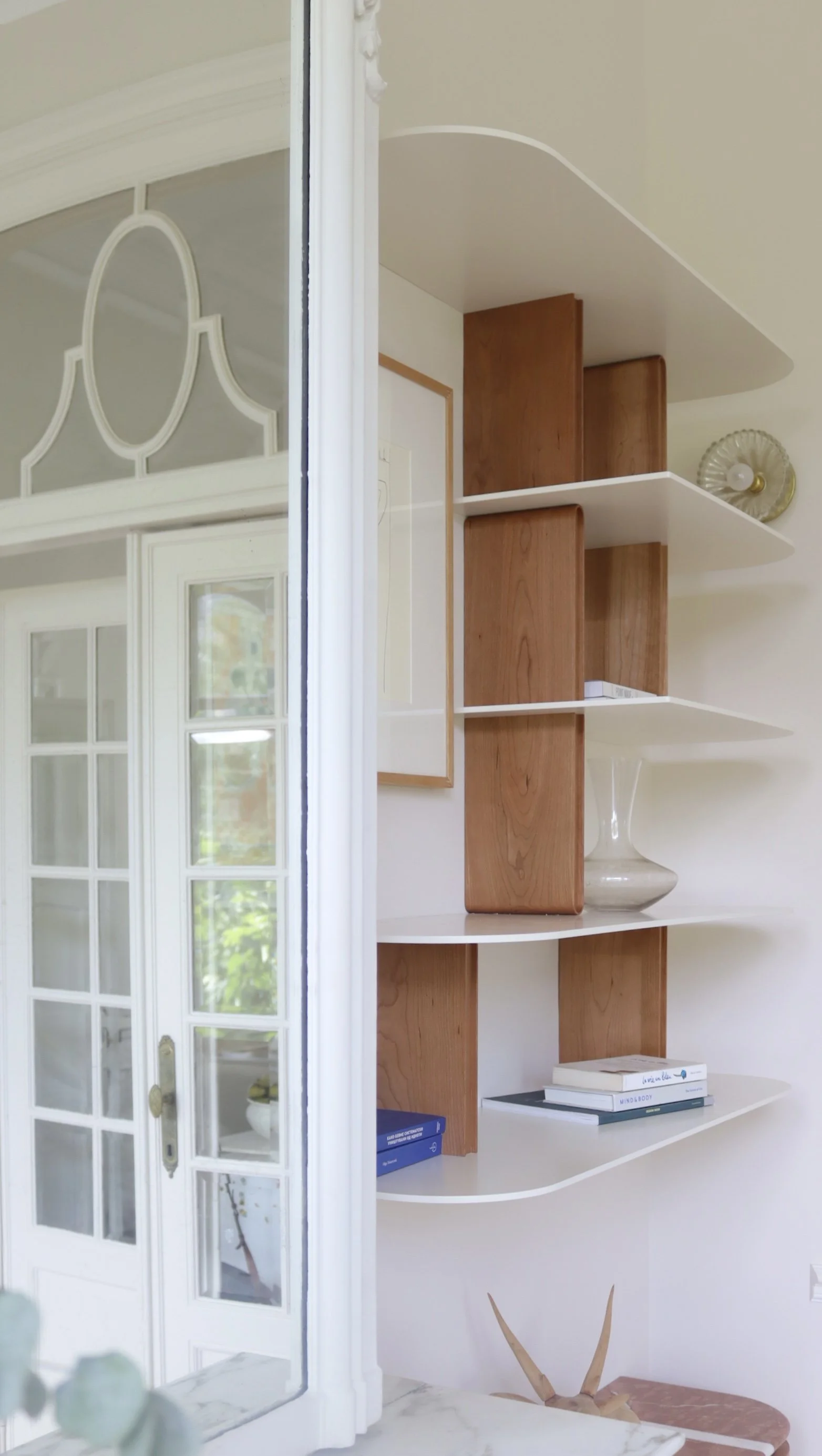 Une partie d'une étagère en bois blanche avec livres, une petite plaque en verre et une pièce décorative en forme d'animal en haut. À gauche, une porte-fenêtre en verre avec une grille.