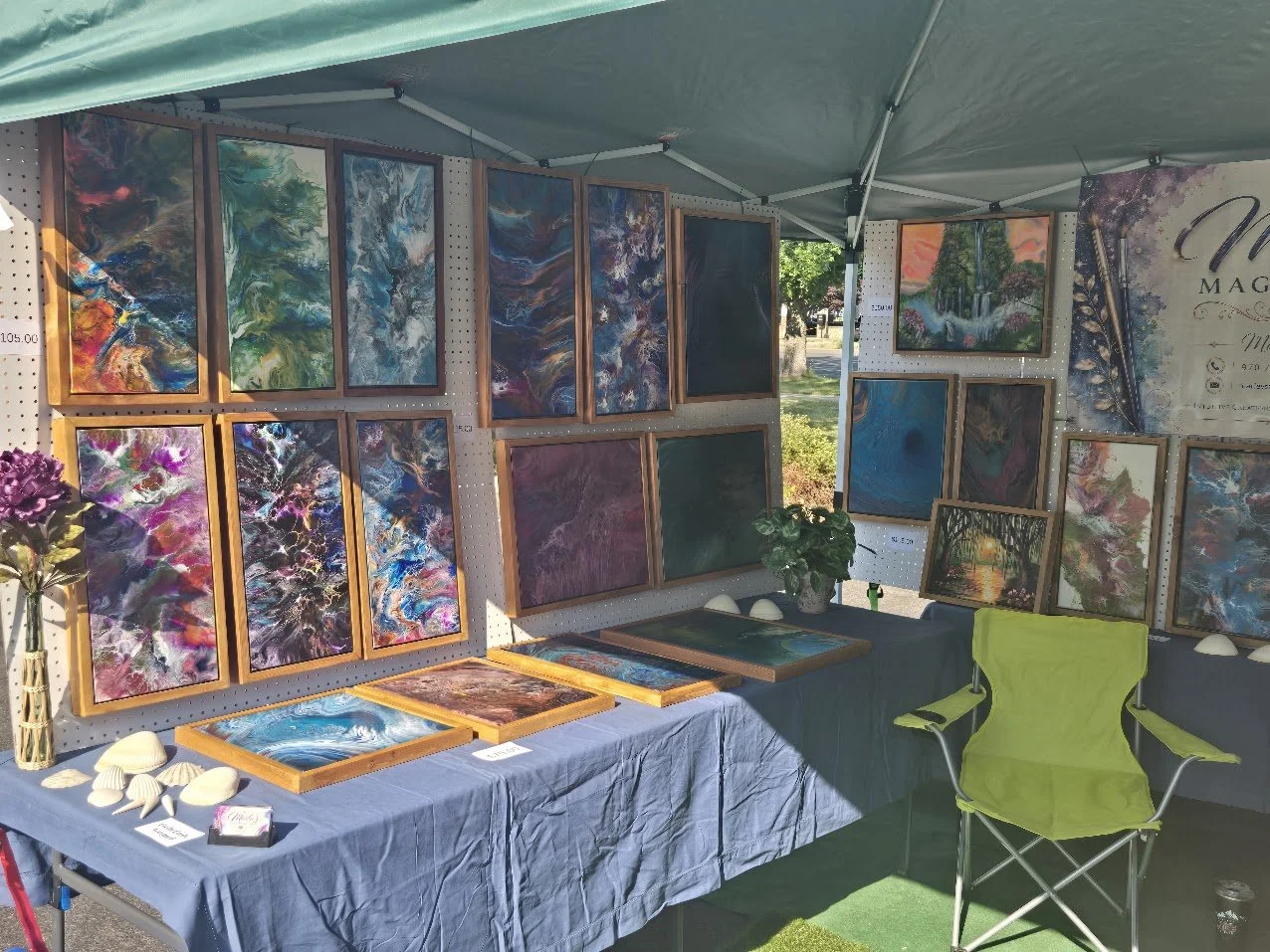 An outdoor art booth displaying colorful abstract paintings in frames on pegboards. A table with shells and a small plant is in front, with a lime green chair beside it.