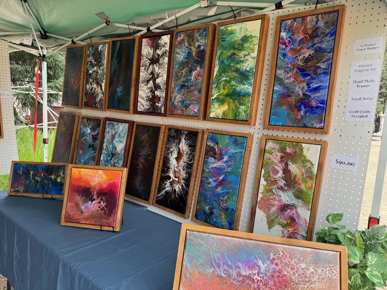 Display of colorful abstract paintings at an outdoor art booth, with paintings framed in wood and arranged on a pegboard wall and on a table.