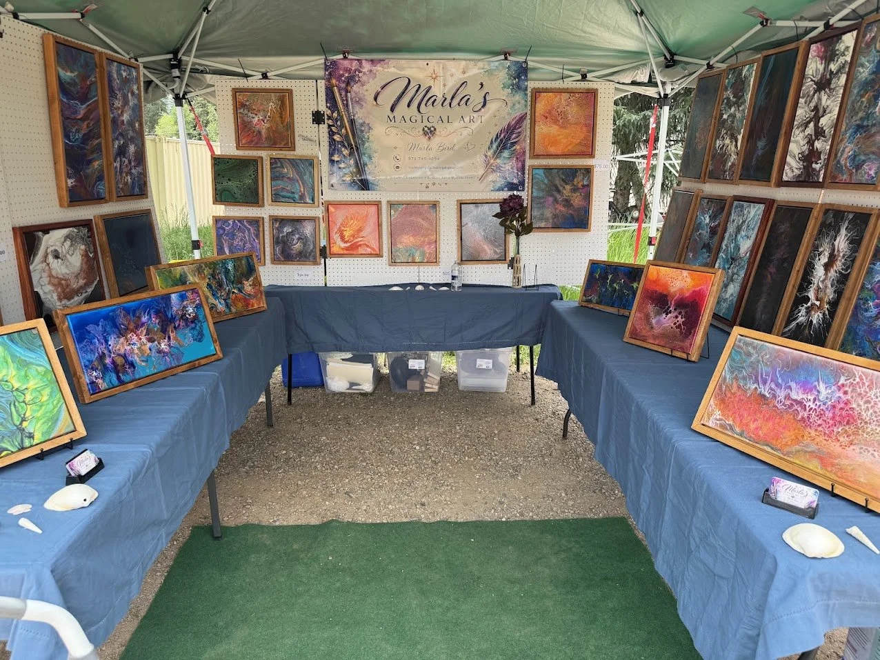 An art display booth showcasing colorful abstract paintings on framed canvases, arranged on tables and pegboards, with a sign reading 'Marla's Magical Art' at the top center.