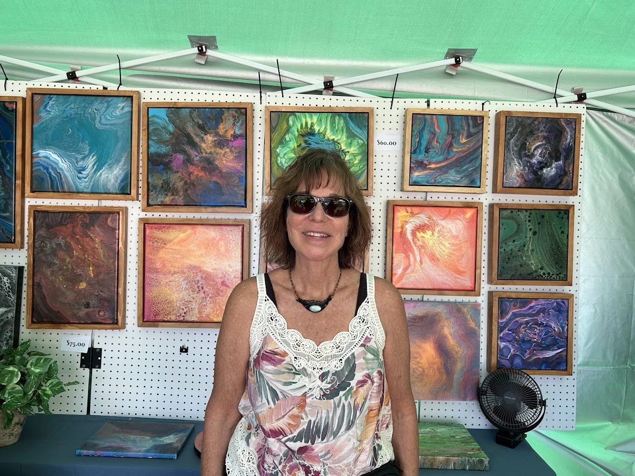 A woman wearing sunglasses and a floral top standing in front of an art display at an outdoor art fair, with colorful abstract paintings behind her.