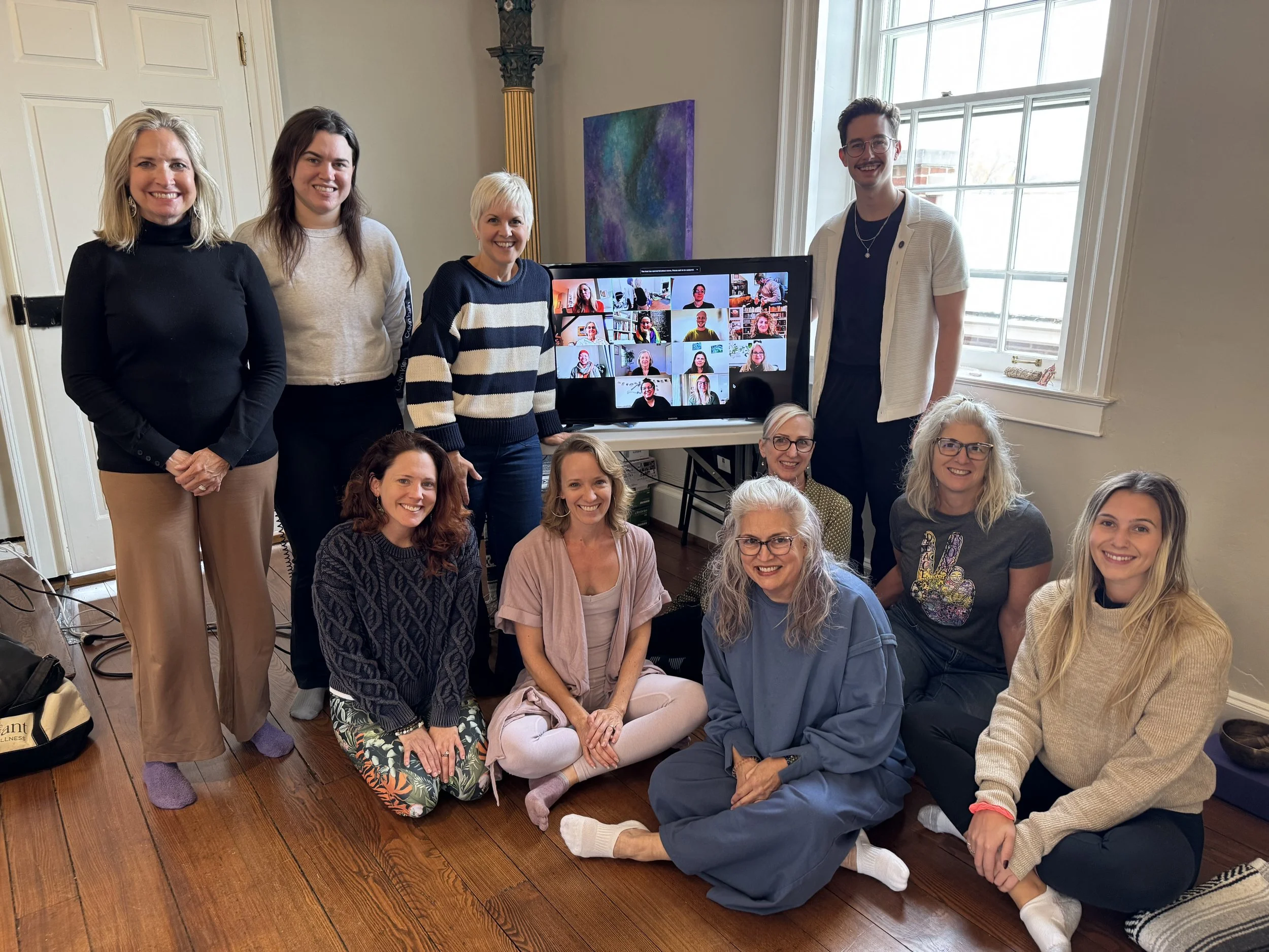 Group of women and men gathered in a room with a large window, some standing and others sitting on the floor, with a monitor showing images of people on a virtual call for an iRest Level one Training.
