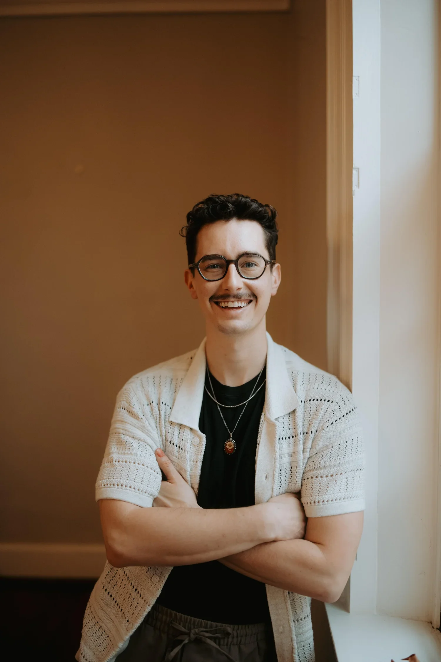 A smiling young man with glasses, wearing a light-colored knit cardigan over a black shirt, with crossed arms, standing indoors in front of a brown wall and a white door.