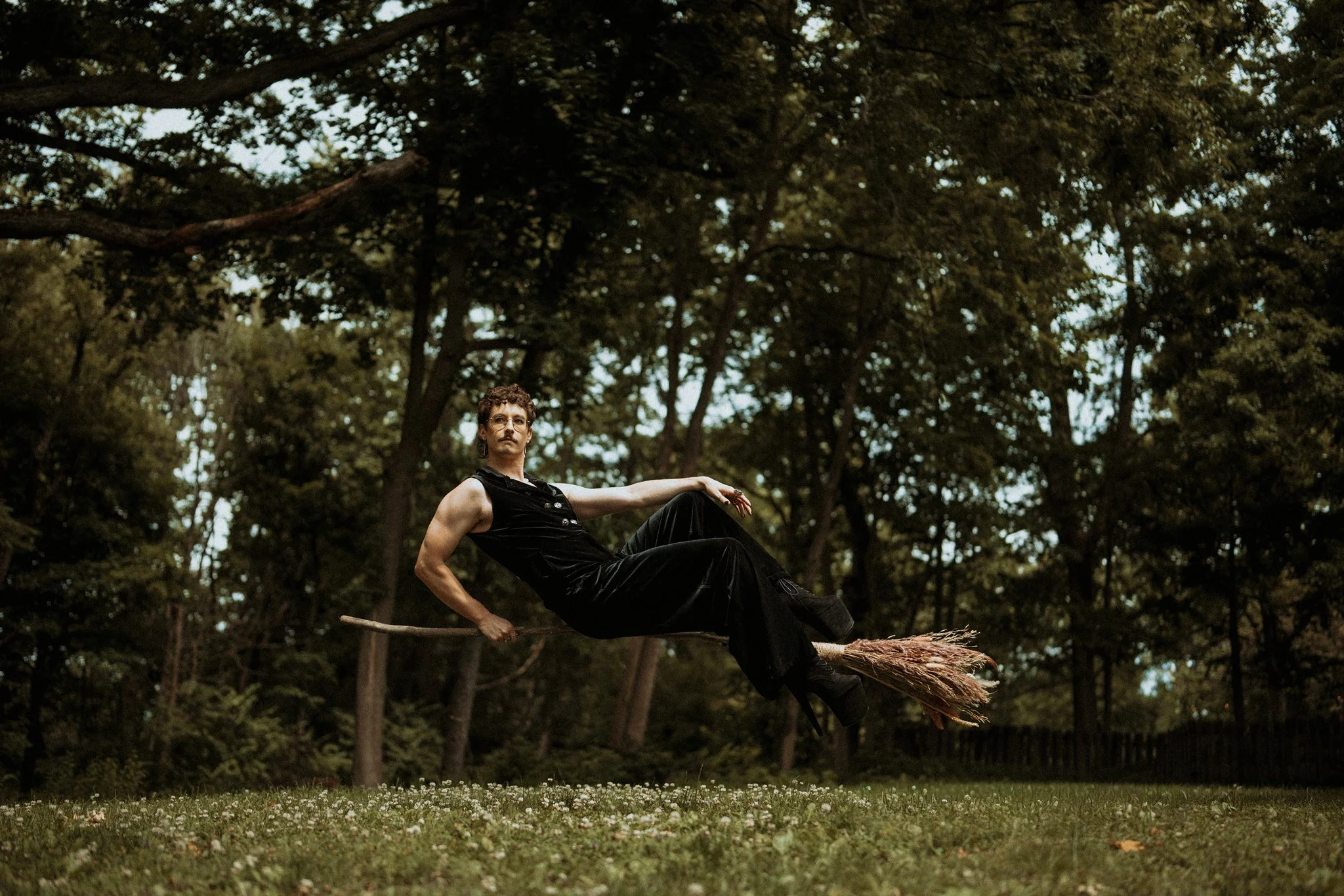 A person with curly hair and glasses sitting on a broomstick in a forested area, with a dense canopy of tall trees overhead and a grassy ground with small white flowers.