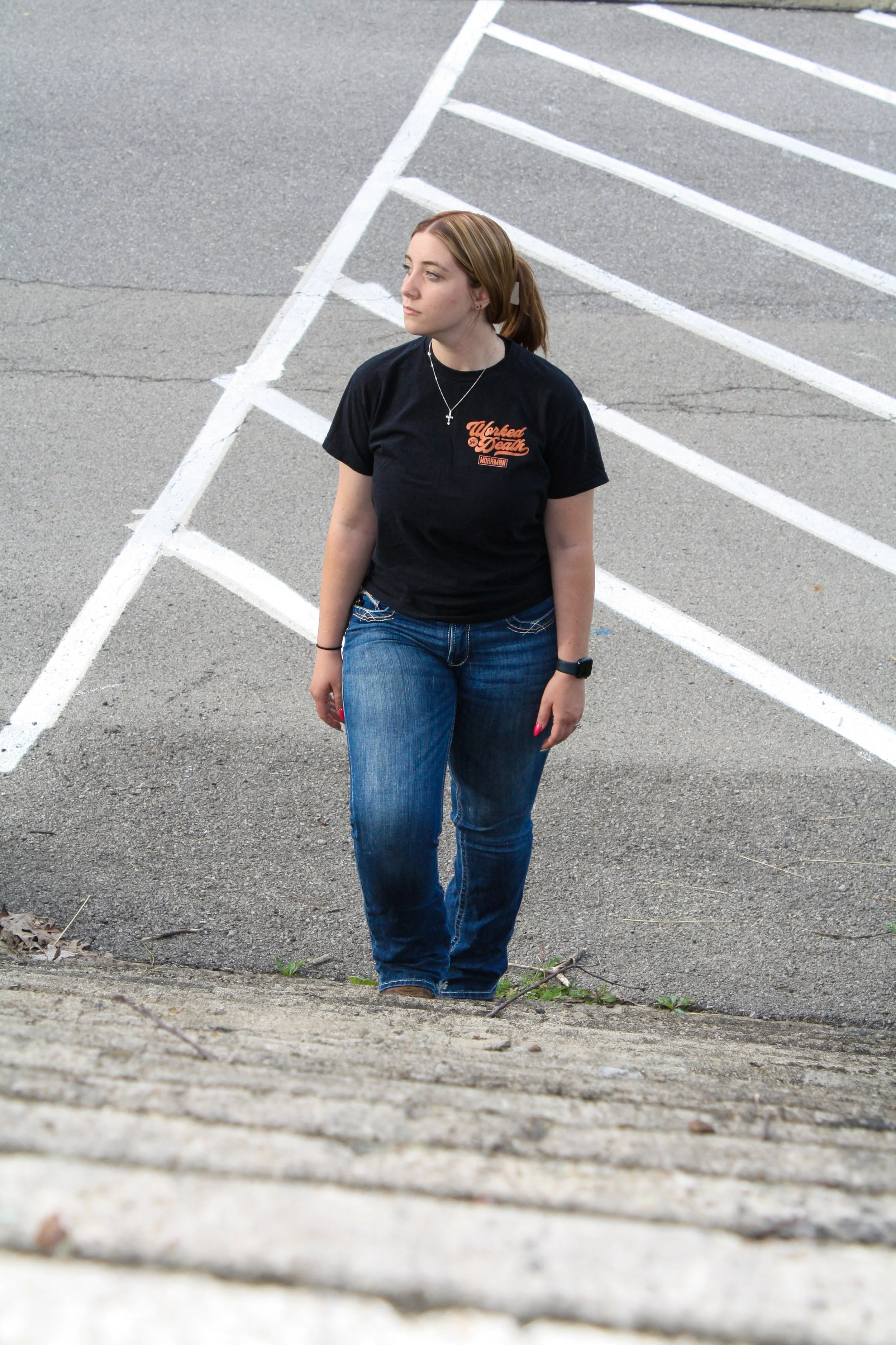 A young woman with red hair in a ponytail, wearing a black T-shirt with orange and white text, blue jeans, and a bracelet, stands outside on a sidewalk with parking lot lines and asphalt visible.