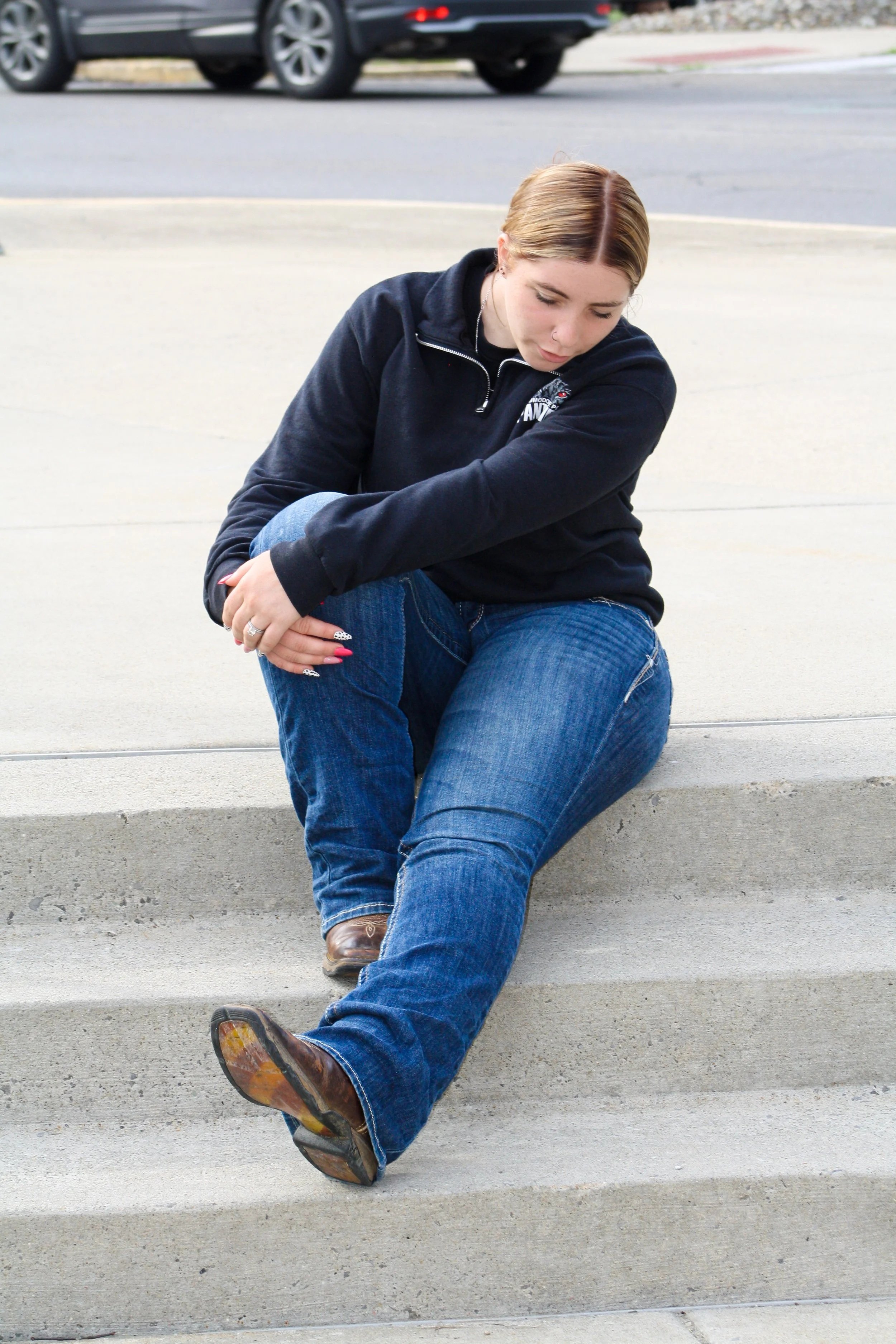 A woman sitting on concrete stairs with her right knee pulled up and her left arm wrapped around it, looking down with closed eyes. She has blonde hair tied back and is dressed in a black zip-up hoodie, jeans, and brown boots. A black car is visible 