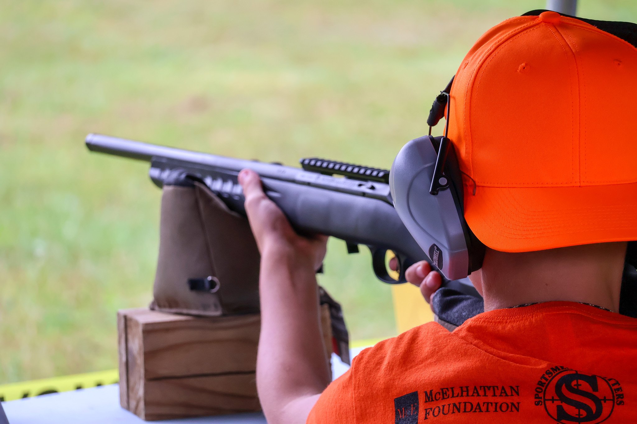 A person wearing an orange shirt and a bright orange cap, aiming a rifle at a target outdoors, with a wooden stand in front of them.