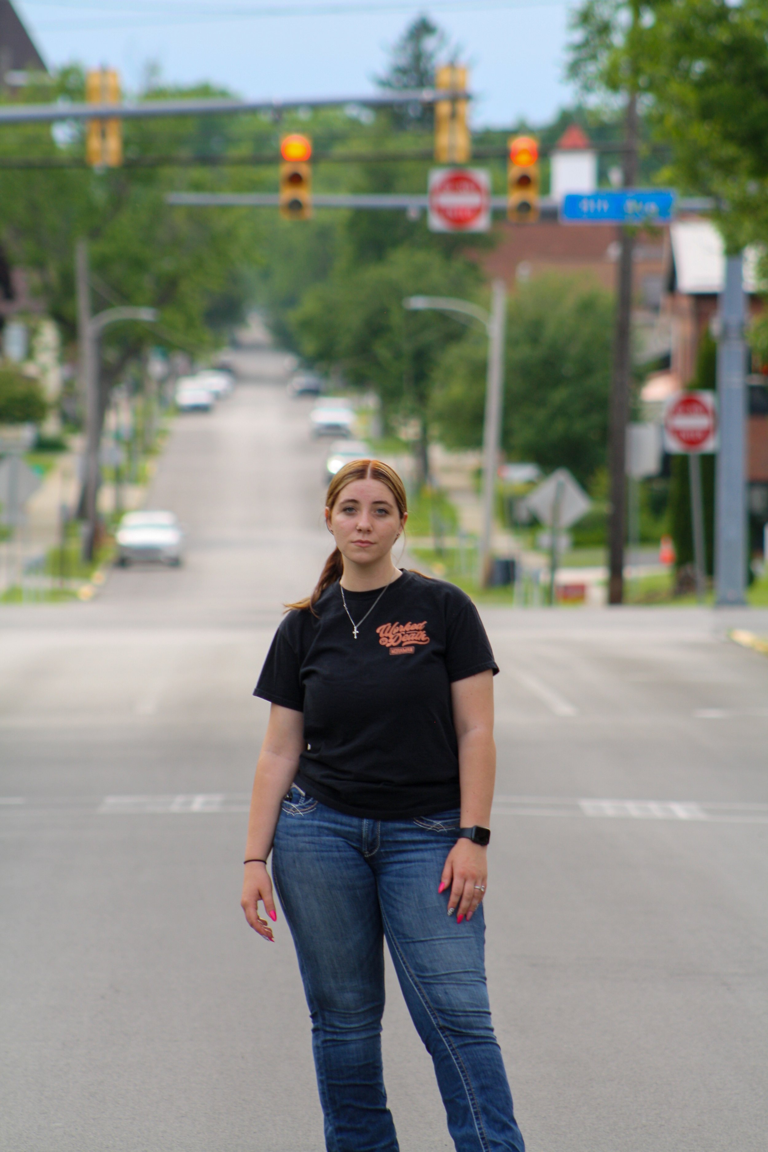A young woman with brown hair tied back, wearing a black t-shirt and blue jeans, standing in the middle of a parking lot during daytime, with a blurred street and traffic lights in the background.