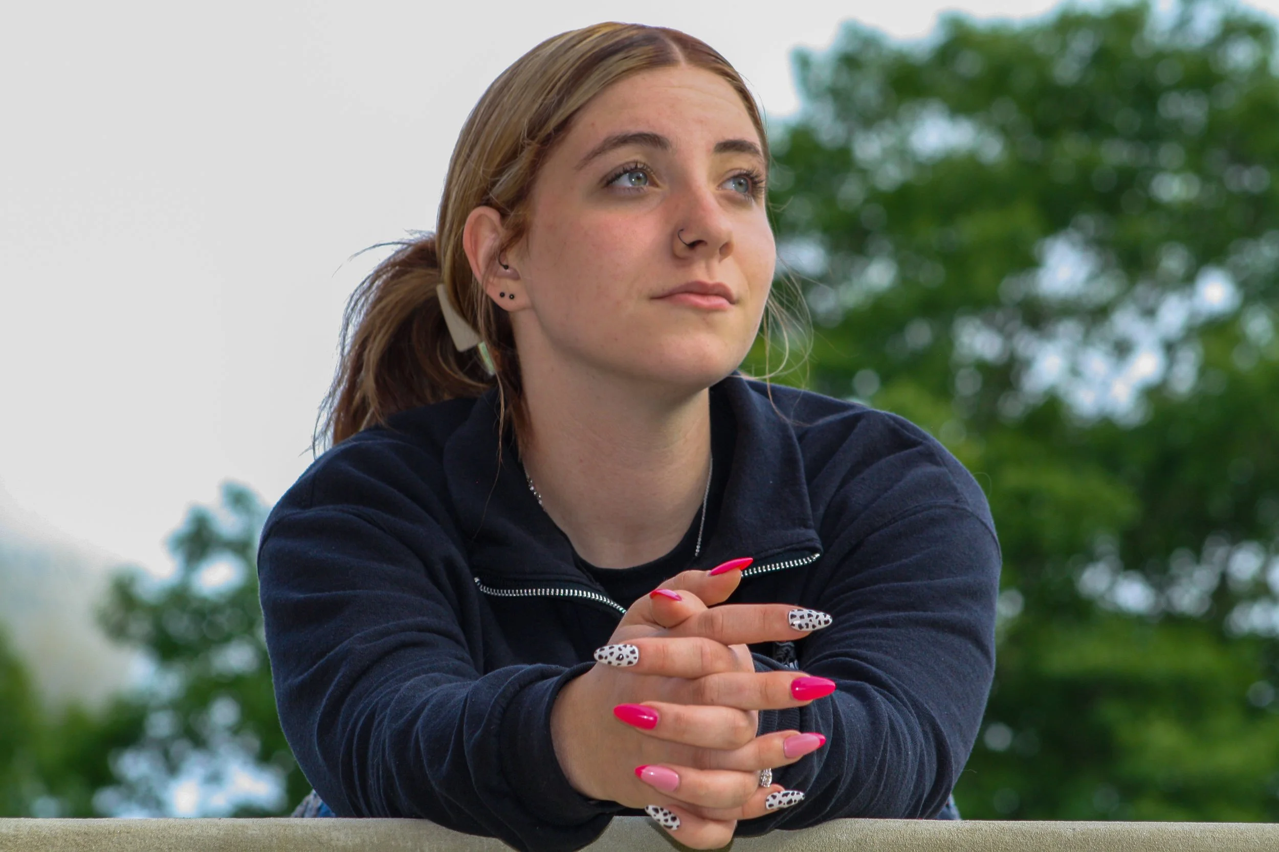 A young woman with red hair, blue eyes, and multiple earrings is sitting outdoors with a background of trees. She is wearing a dark jacket and has decorated nails with various designs, including leopard print and colored polish. She appears to be loo