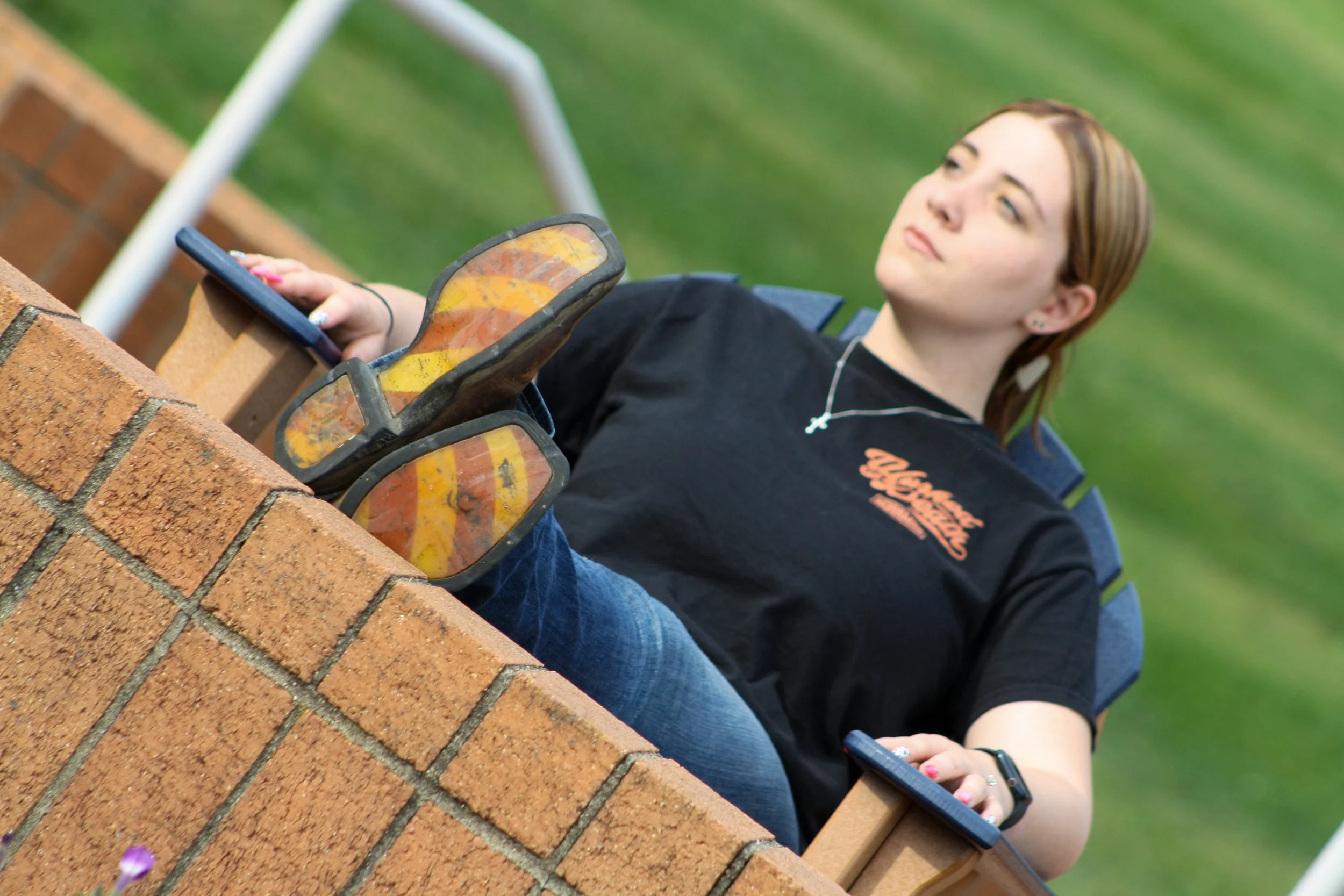 A young woman with brown hair, wearing a black t-shirt and a necklace, sitting outdoors on a chair, holding a smartphone, with a brick wall in the foreground and green grass in the background.