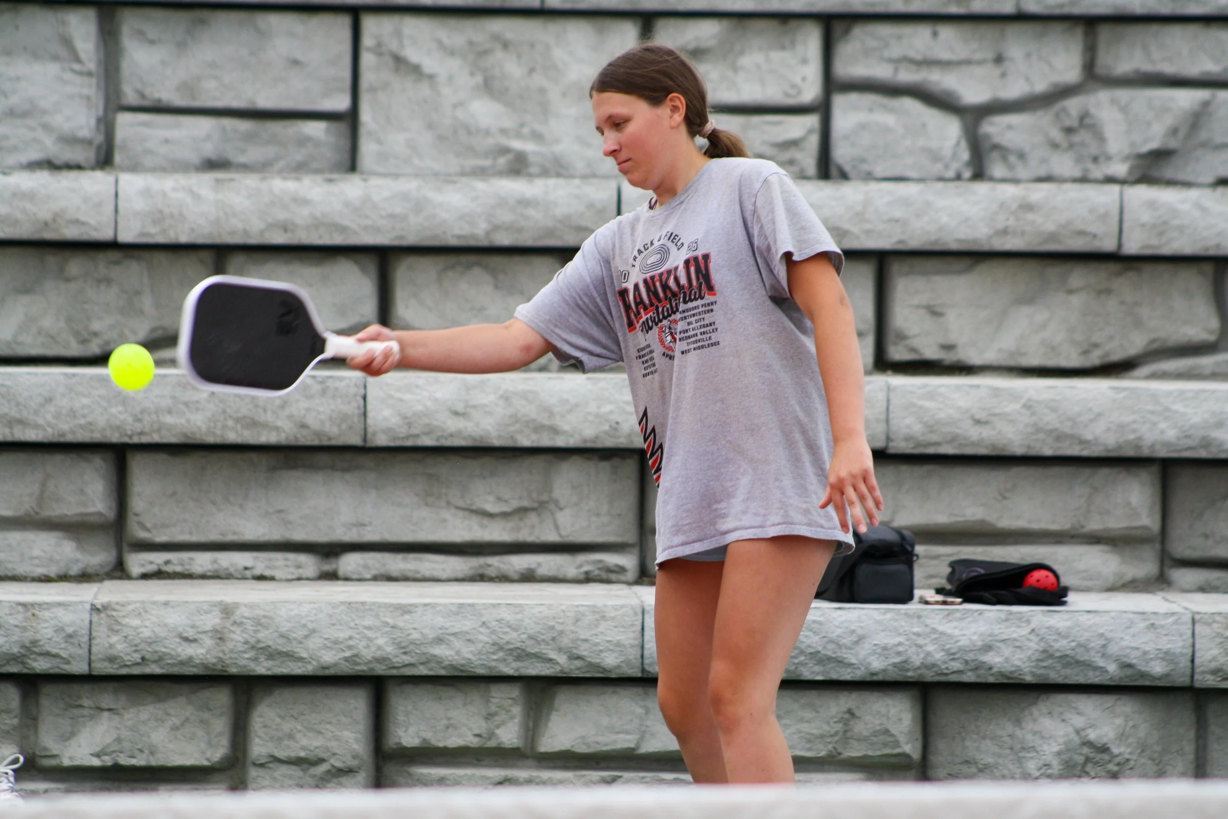 A girl in a gray T-shirt and shorts playing pickleball outdoors on a set of gray stone stairs. She is holding a pickleball paddle and about to hit a yellow pickleball.