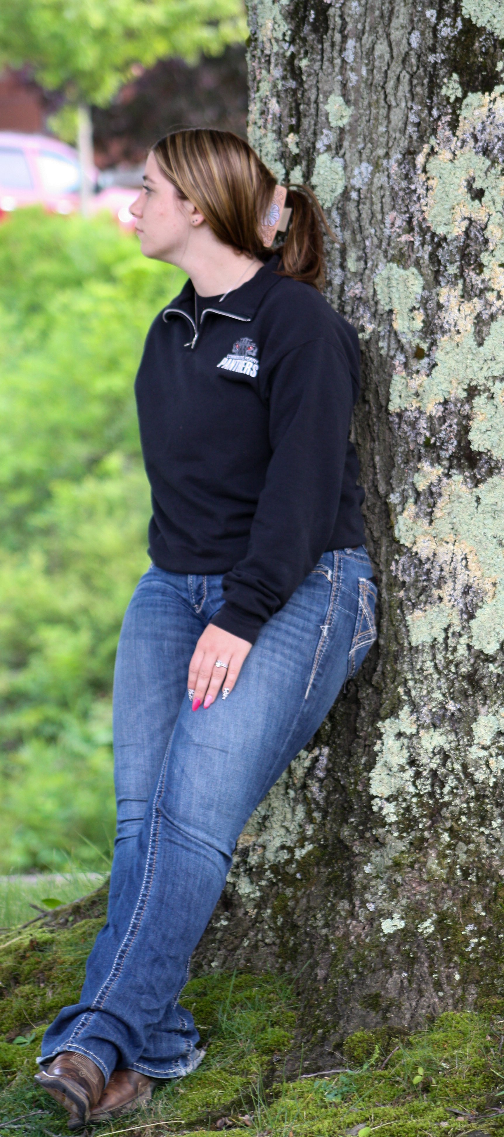 A woman with brown hair in a ponytail, wearing a black sweatshirt and blue jeans, standing outdoors and leaning against a tree trunk with moss and lichen.