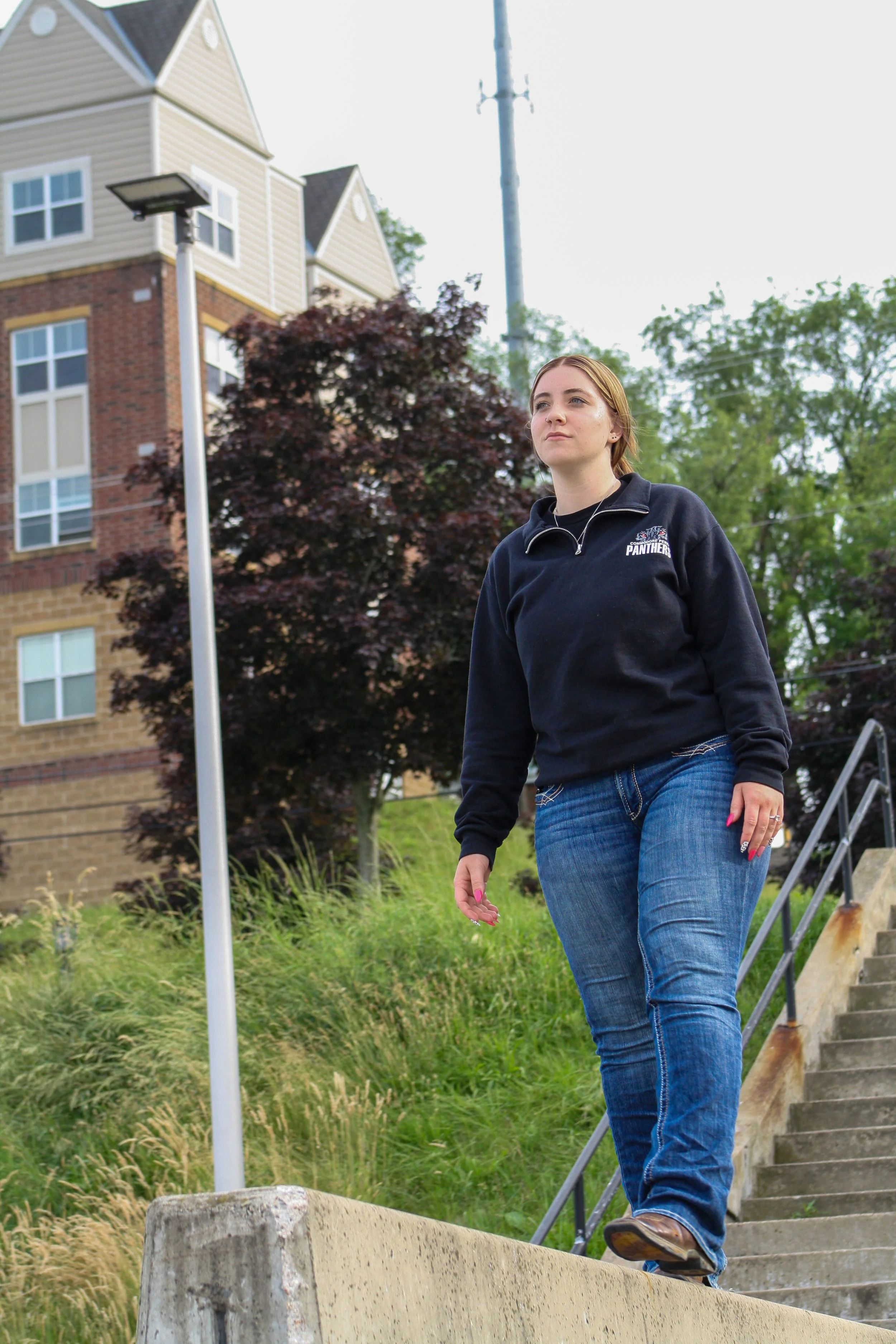 A young woman with reddish hair wearing a black sweatshirt with a sports team logo, blue jeans, and brown shoes walking down outdoor concrete stairs near a grassy area with trees, a flagpole, and a large apartment building in the background.