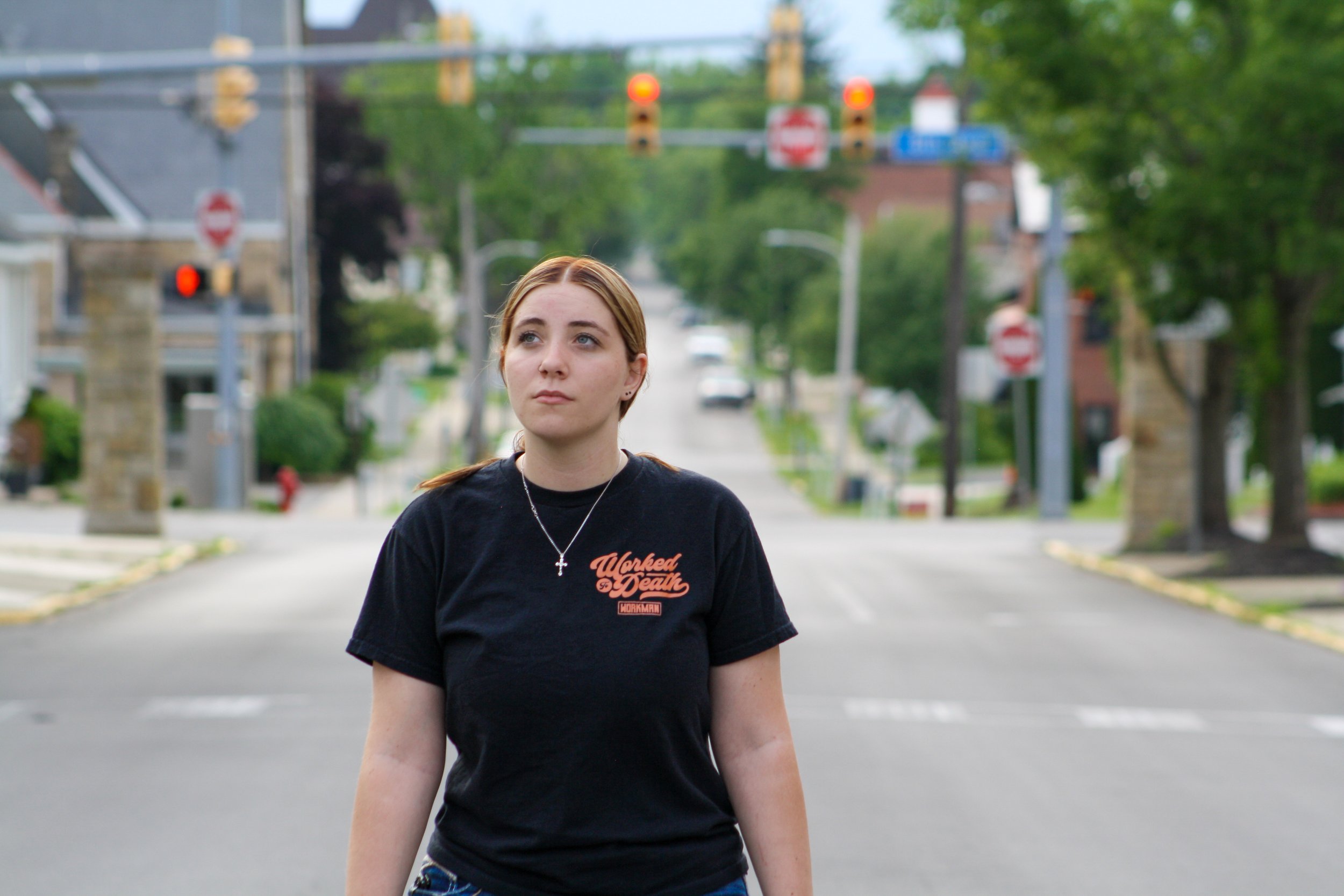 A young woman with brown hair and light skin standing in the middle of a street, looking to her right. She is wearing a black T-shirt with orange text and a silver cross necklace. Behind her, blurred traffic lights and trees line the street.