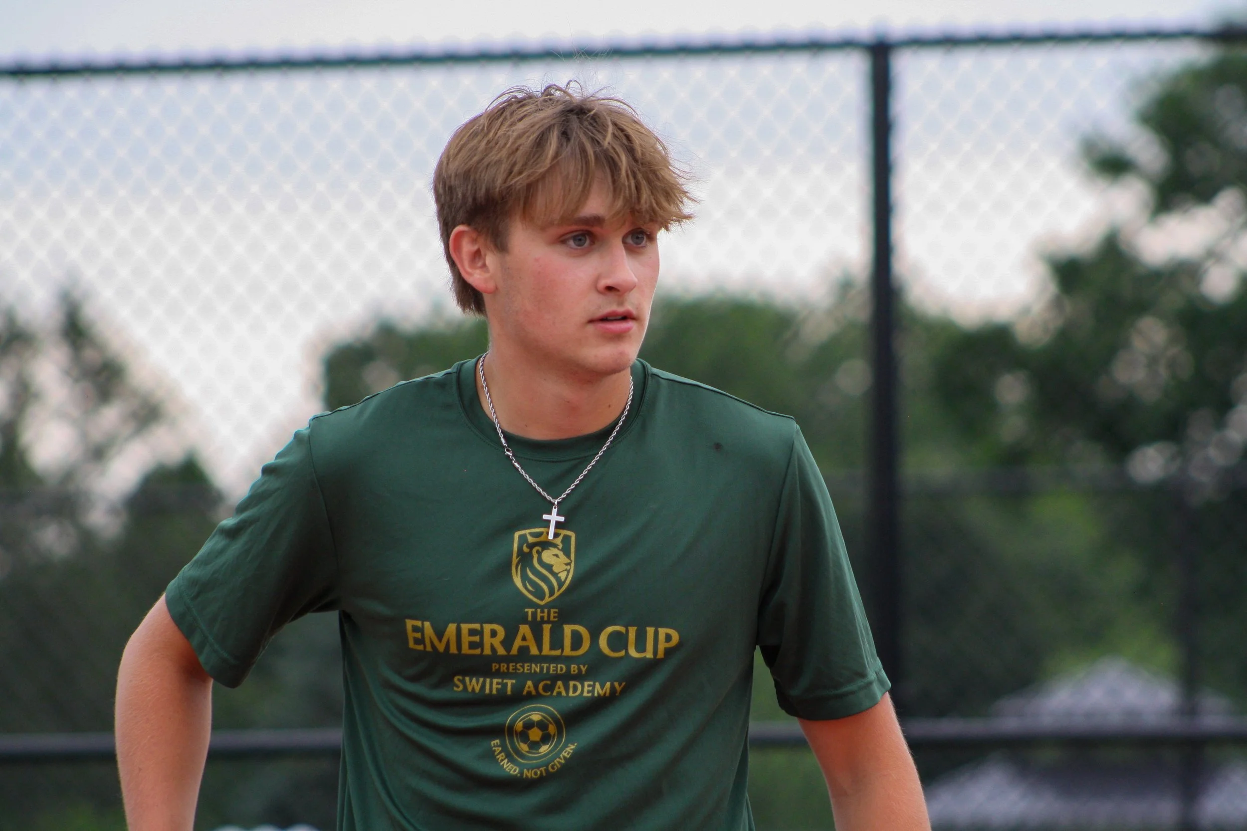 A teenage boy with light skin and tousled blond hair wearing a dark green t-shirt with a soccer emblem and the words 'The Emerald Cup presented by Swift Academy'. He is outdoors on a sports field with a chain-link fence and trees in the background, l