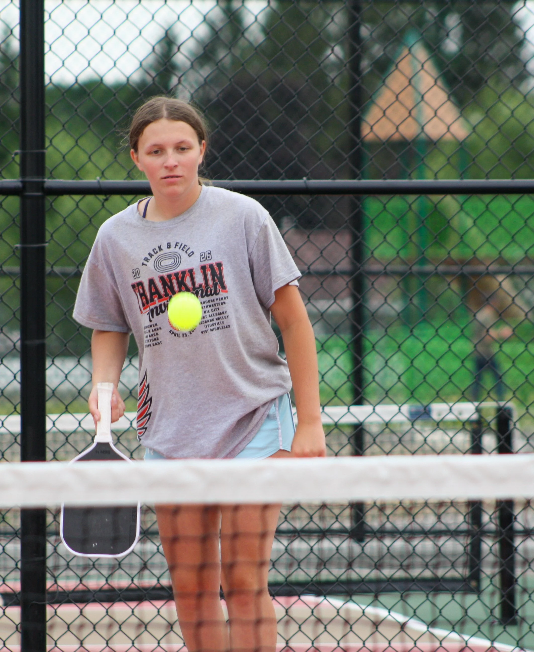 Young woman playing pickleball on an outdoor court, wearing a gray 'Franklin' T-shirt and holding a pickleball paddle, behind a black chain-link fence.
