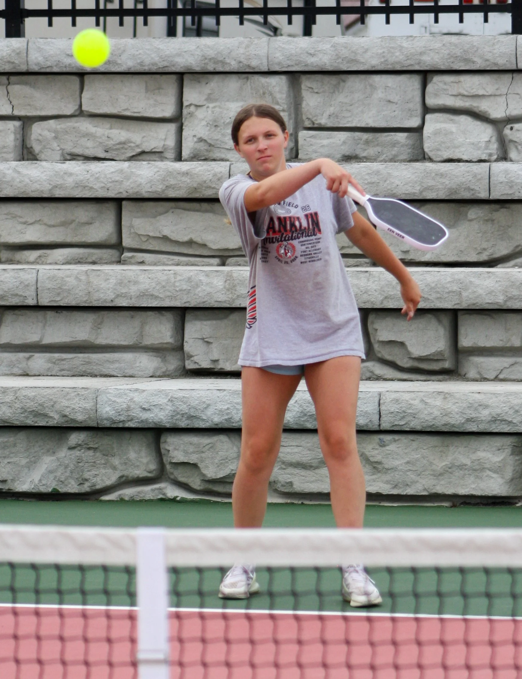A young woman on a tennis court hitting a tennis ball with her racket. She is standing behind a net with a stone wall as the background.