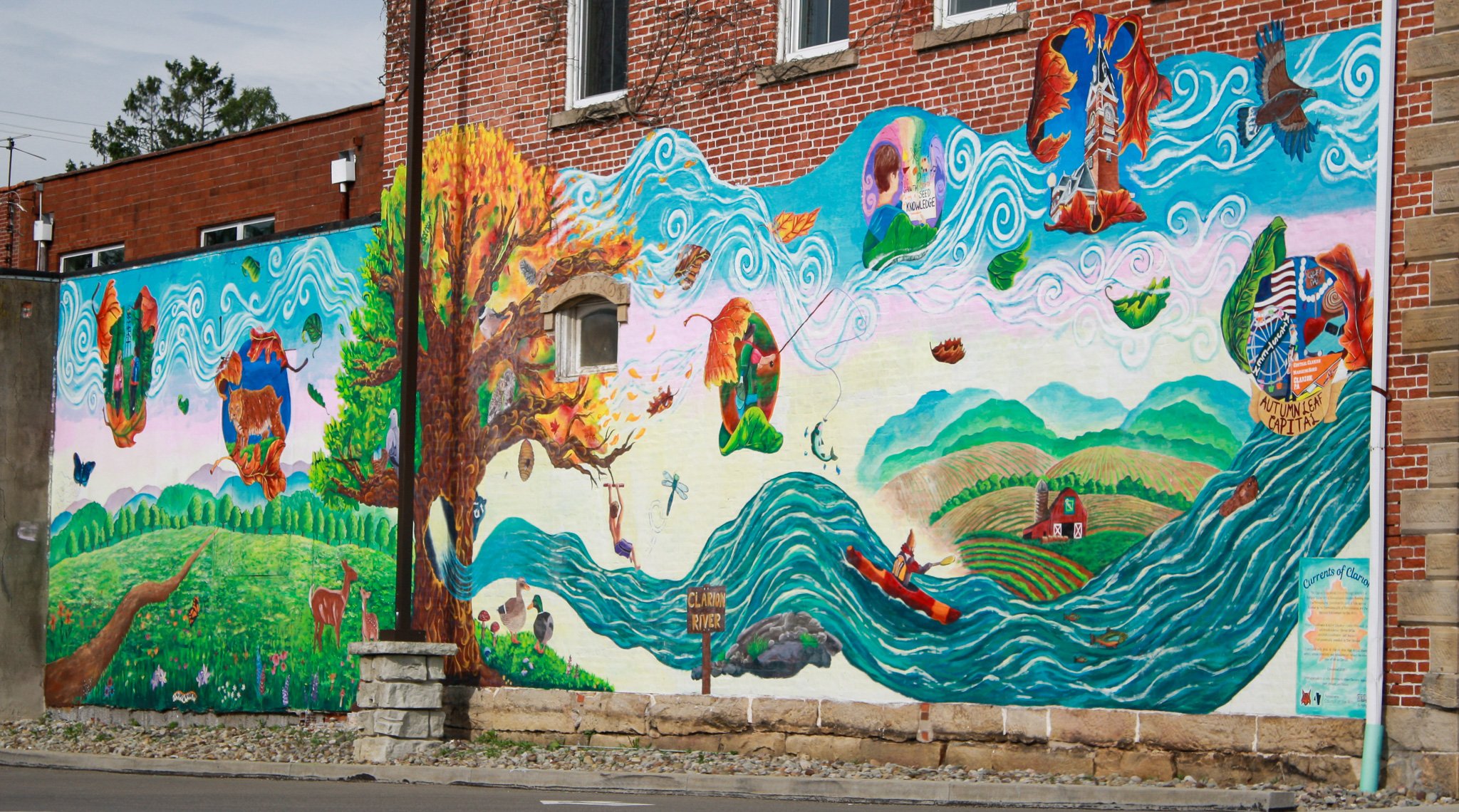 Colorful mural on a red brick building depicting a natural landscape with water, trees, animals, and people engaging in outdoor activities like fishing and kayaking.