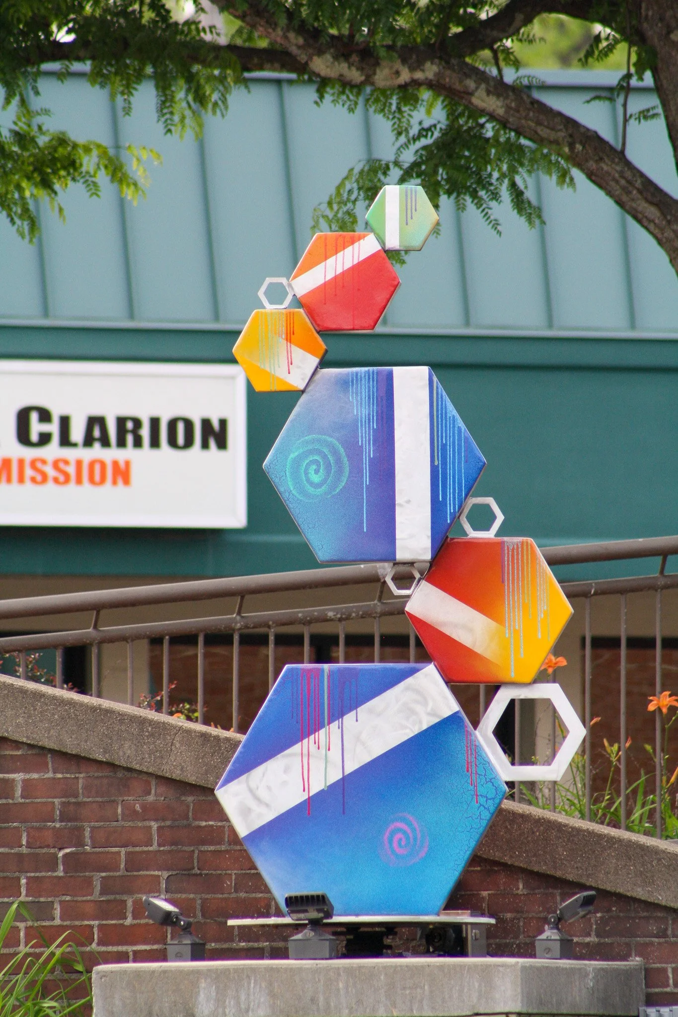 Colorful geometric sculptures with hexagon shapes stacked in a vertical line outdoors, with a brick wall, tree branches, and a sign that reads 'Clarion Mission' in the background.