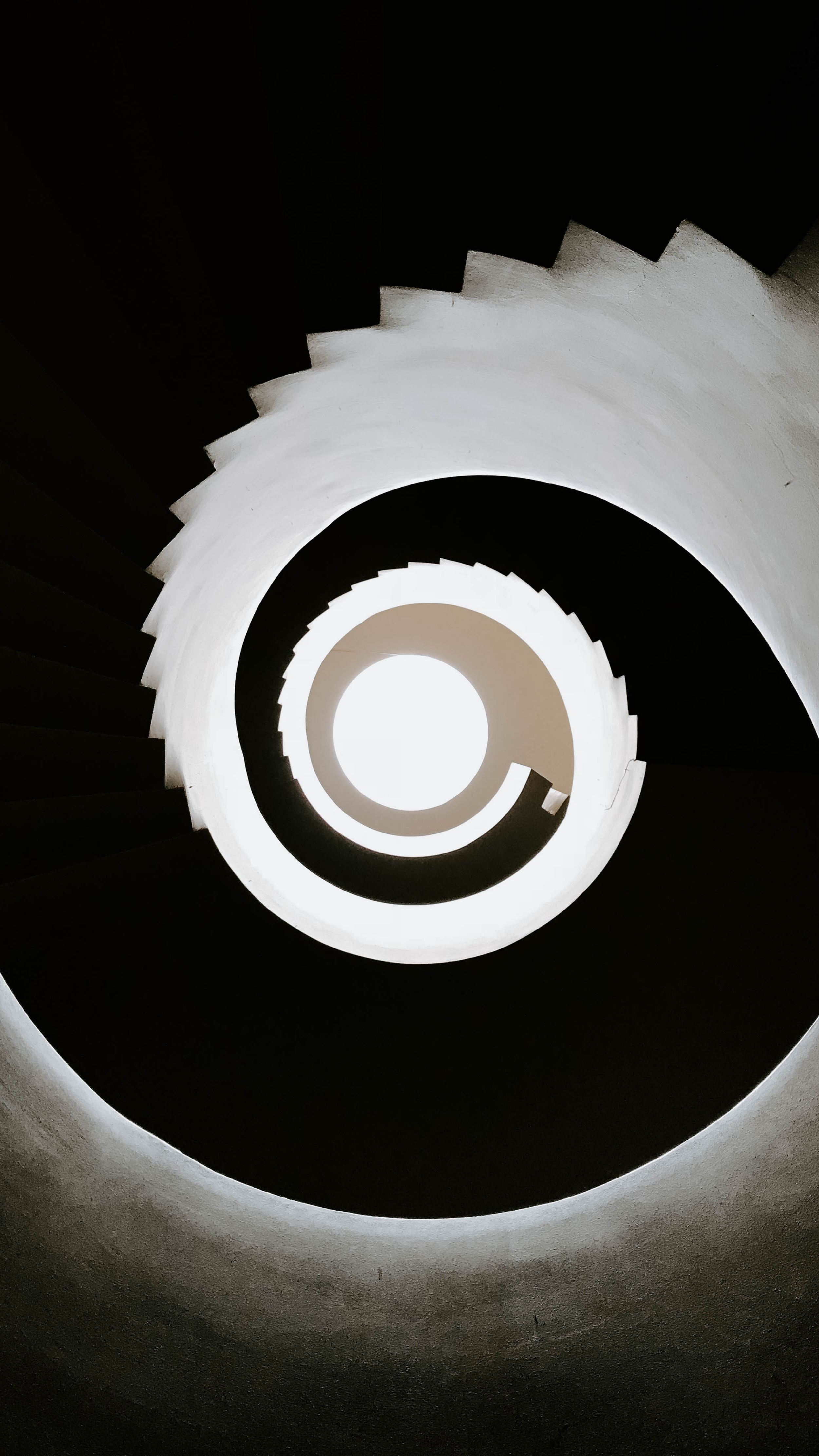 Blick nach oben in einen spiralförmigen Treppenaufgang mit weißen Wänden und schwarzen Stufen, die um eine helle zentrale Öffnung herum angeordnet sind. Sinngemäß steht sie für Übergang, Veränderung, Licht und Schatten.
