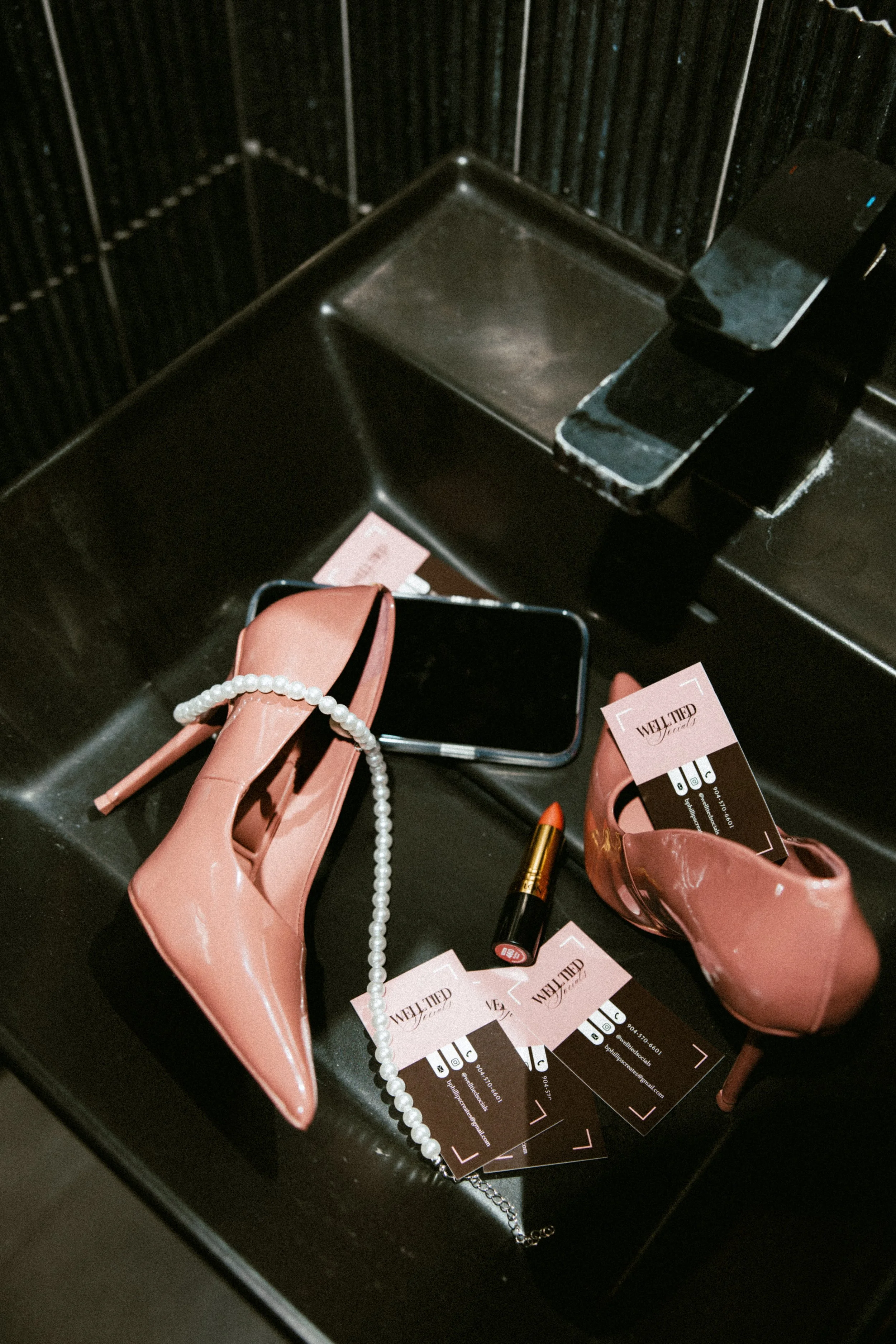 Pink high-heeled shoes with a pearl necklace, business cards, lipstick, and a smartphone on a black surface.