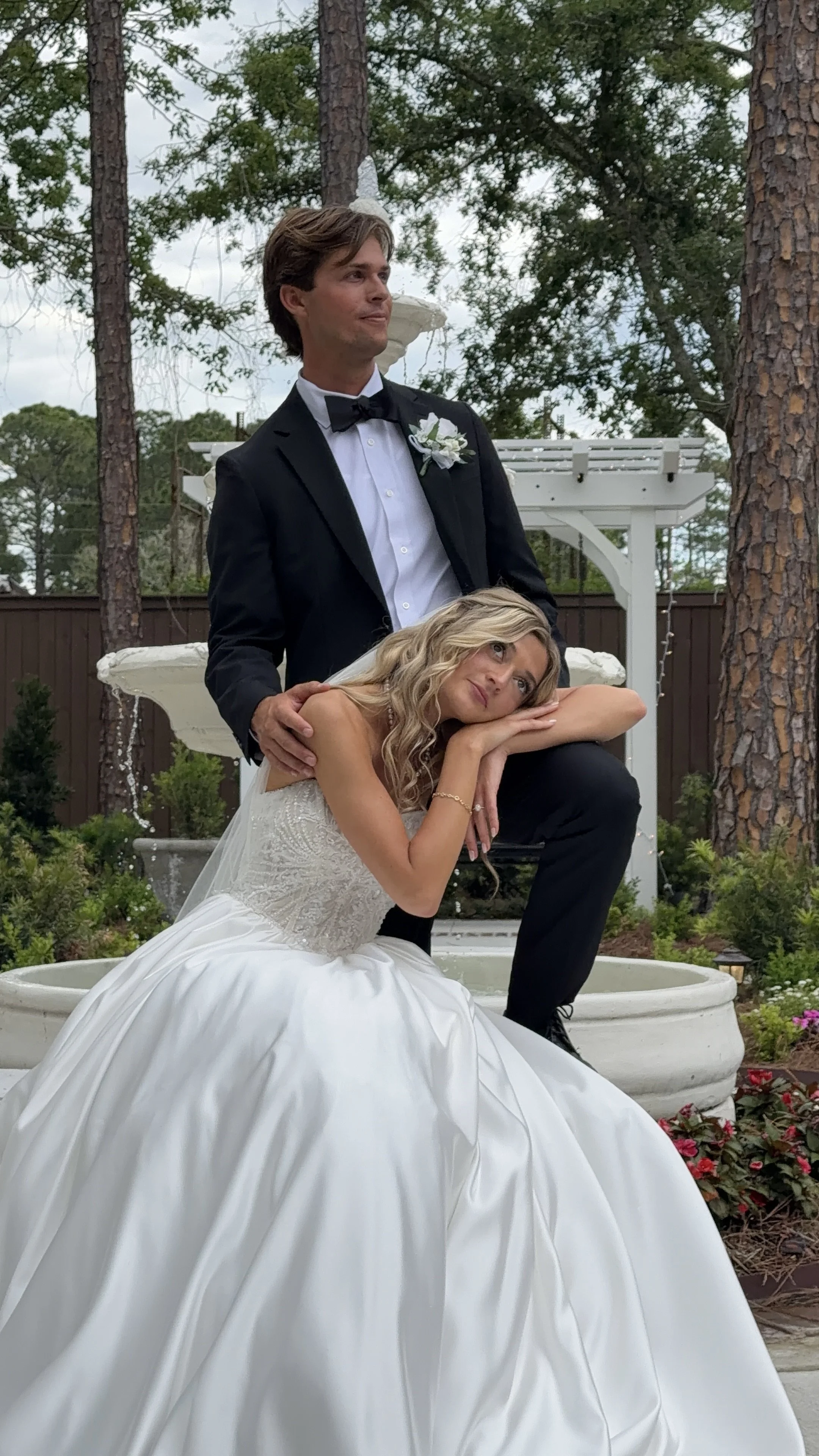 A bride in a white wedding dress sitting in front of a fountain, with a groom in a tuxedo standing behind her and holding her shoulder, in an outdoor garden setting with trees and flowers. Florida Wedding Content Creator