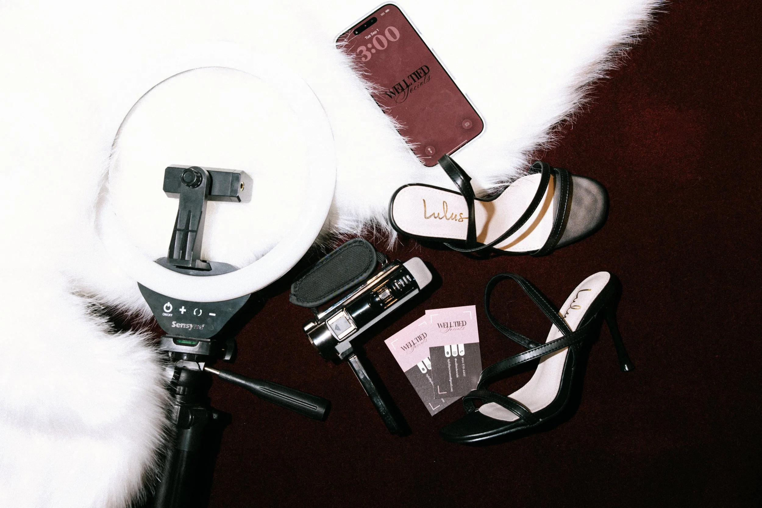 A collection of items placed on a furry white and brown surface, including a white circular device, a smartphone displaying 'WELL FIED Society', a pair of black and white high heel shoes with the label 'Lulus', a black cylindrical object, and promotional cards for 'WELL FIED' products.