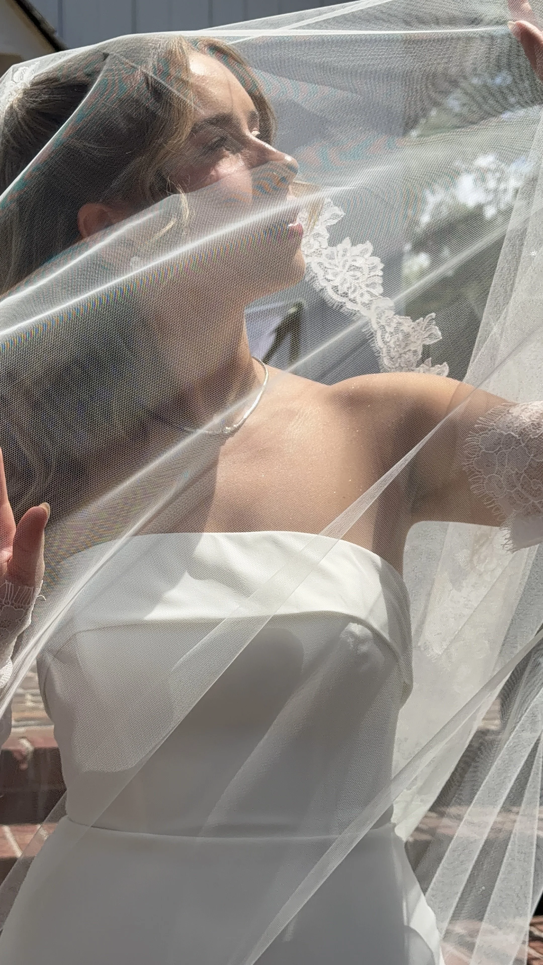 A woman in a white dress is looking through a window with lace curtains, with sunlight shining on her face.