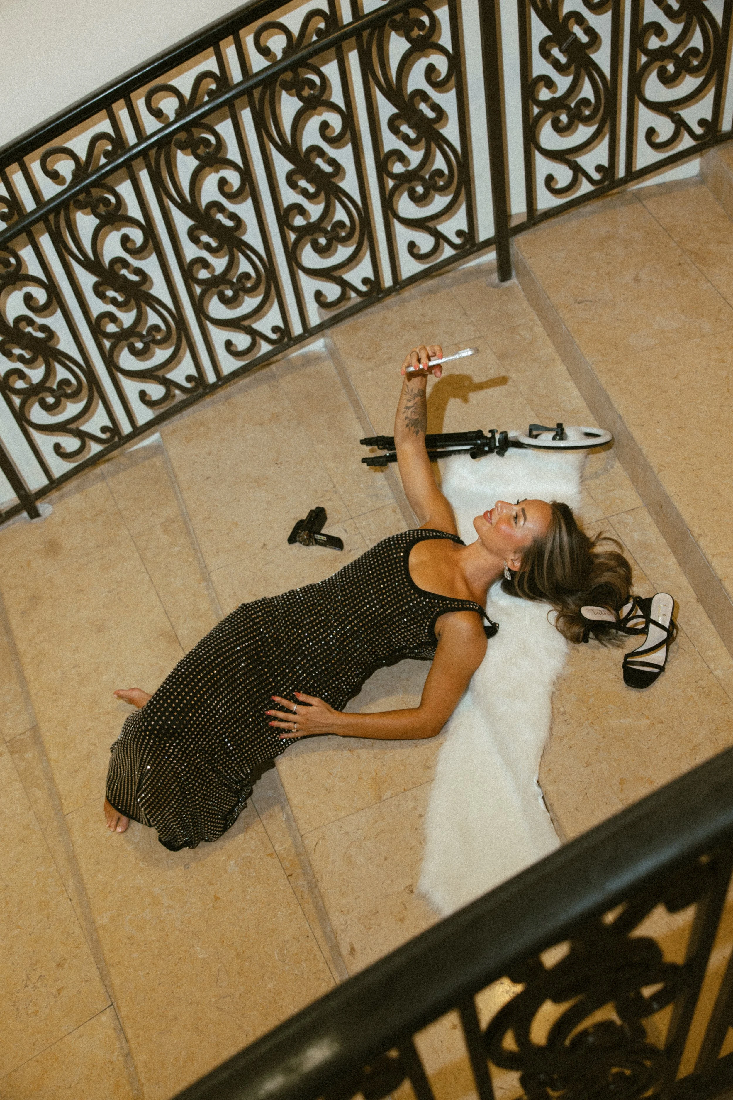 A woman lying on her back on a beige tiled floor, wearing a black and gold dress, holding a cigarette in her right hand, with high-heeled shoes and a white fluffy blanket nearby, and a white scooter in the background.