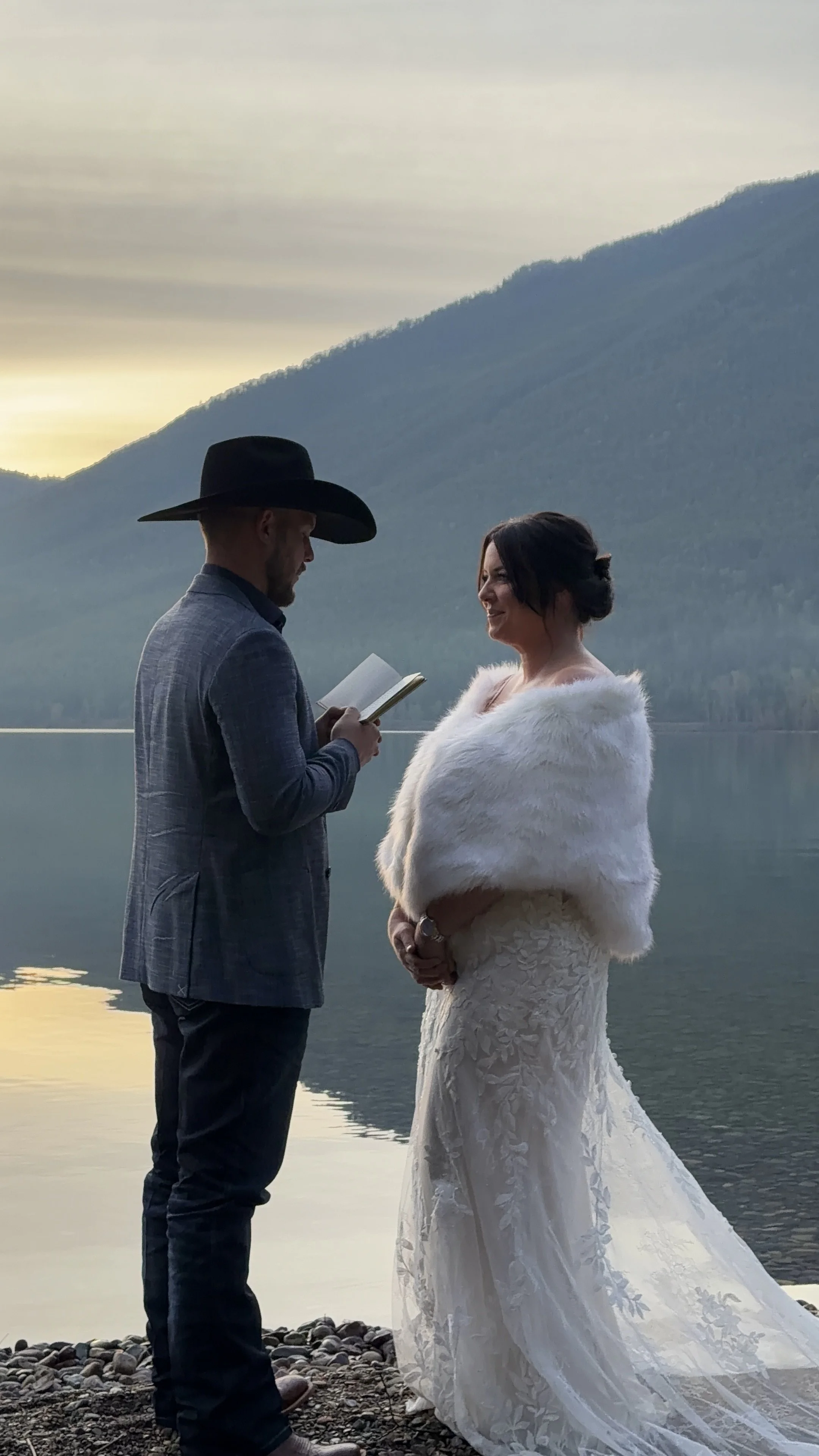 A man and woman standing by a lake during sunset, with mountains in the background. The man is wearing a grey jacket and a black cowboy hat, holding a notebook. The woman is dressed in a white lace wedding gown with a faux fur stole, smiling at the man.