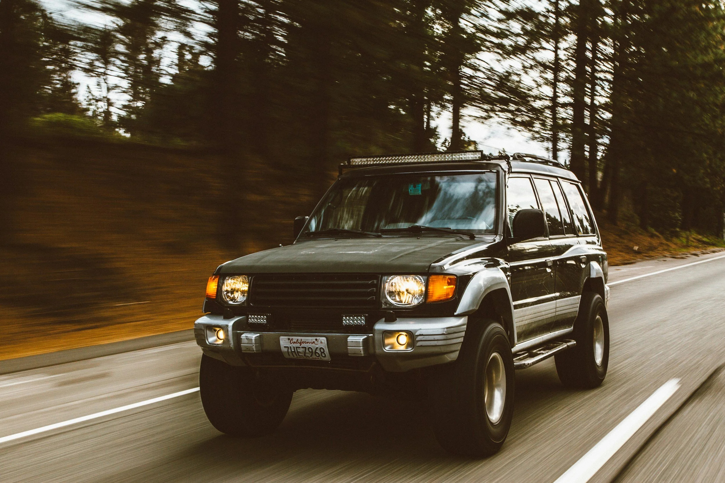 A black SUV driving at speed on a road surrounded by trees, with motion blur indicating movement.