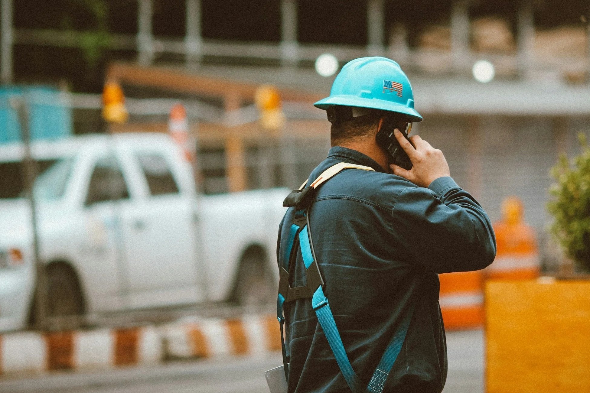 A construction worker wearing a light blue safety helmet with an American flag sticker, talking on a cellphone at a construction site with a white vehicle and construction barriers in the background.