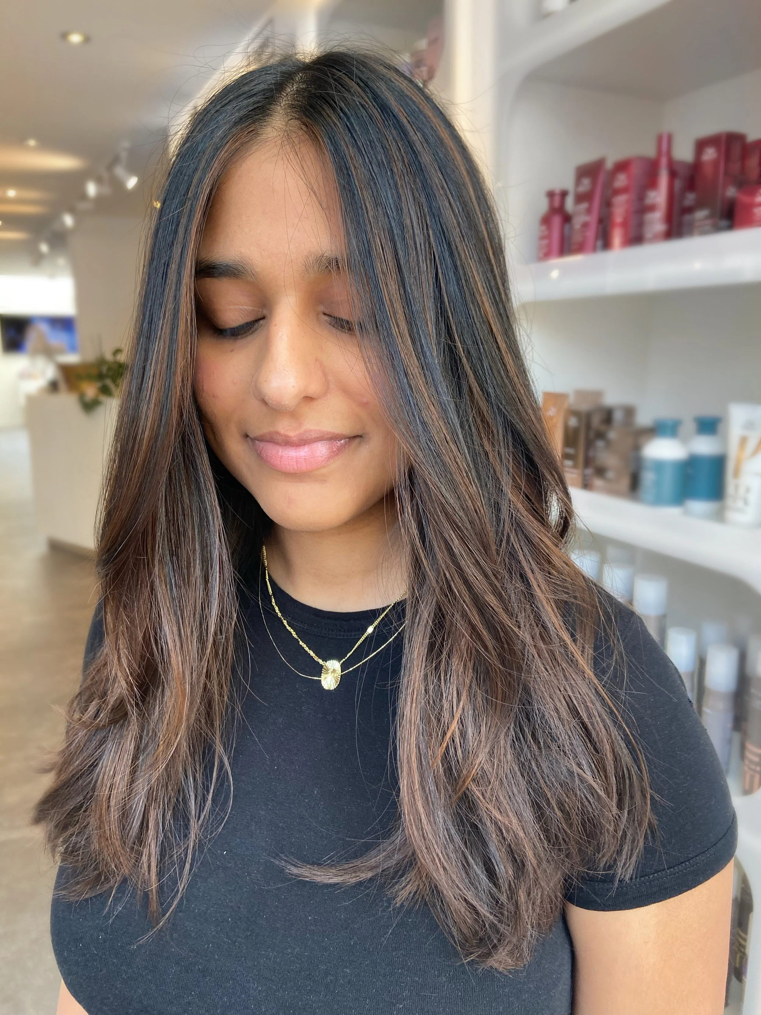 A woman with long, wavy hair dyed with highlights, wearing a black top and layered gold necklaces, standing in a store with shelves of hair products in the background.