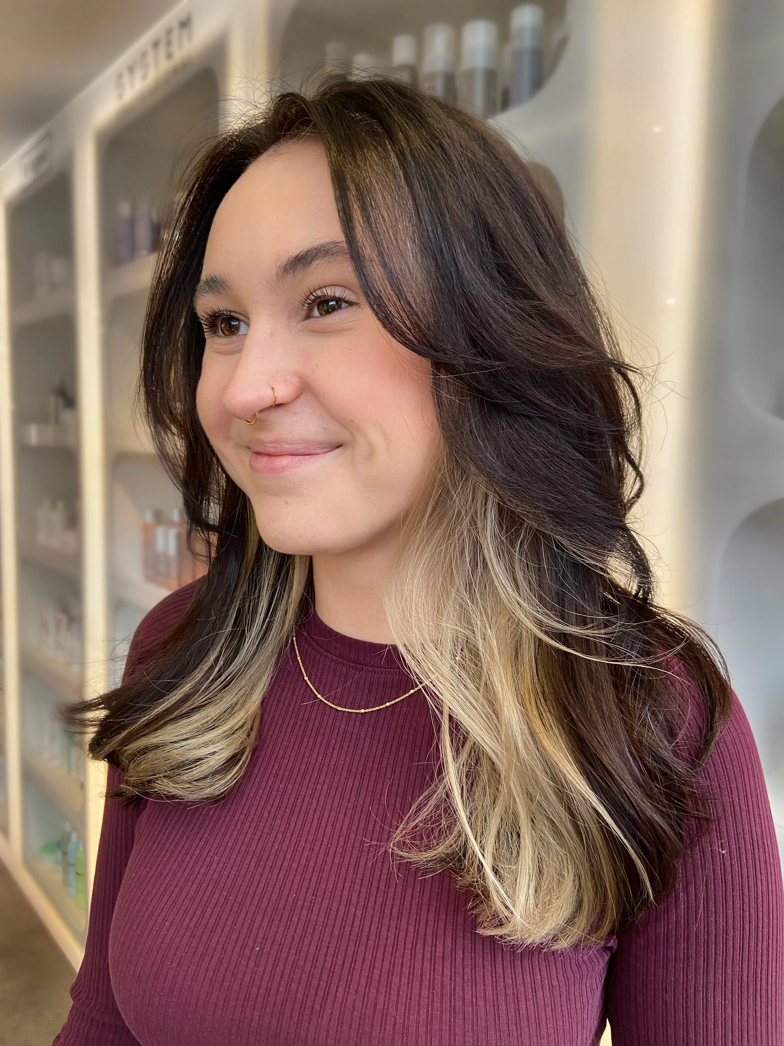 A woman with dark brown hair and blonde block color, wearing a maroon top and gold necklace, is smiling slightly. She has a nose ring and is standing in front of shelves with various hair or skincare products in a salon or store.