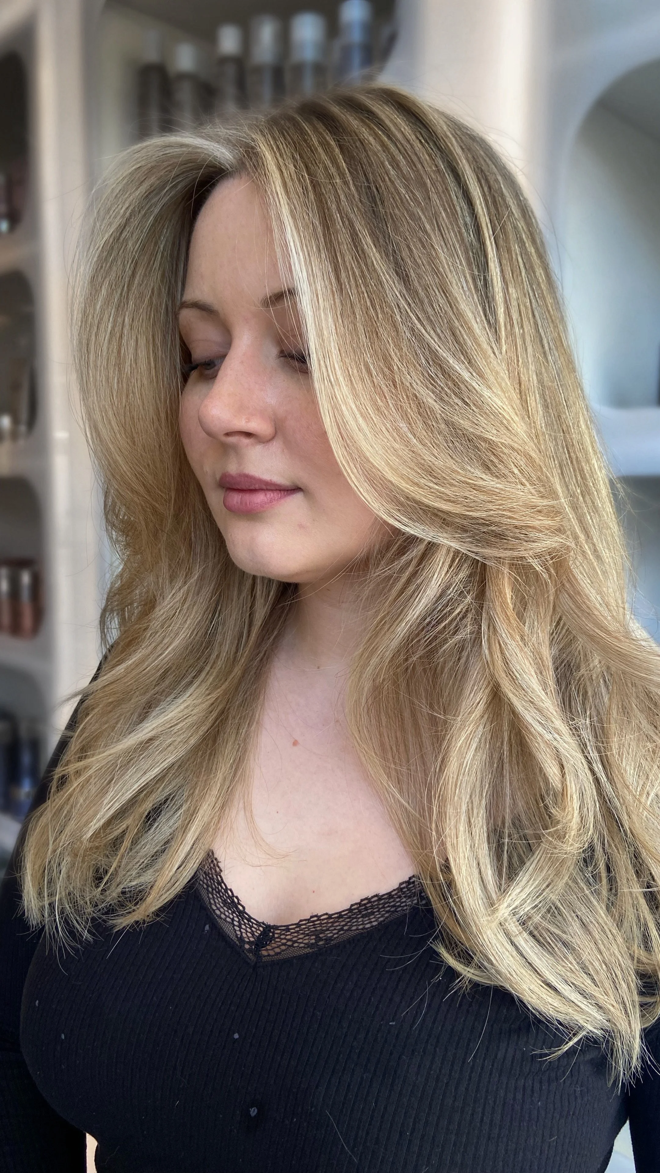 A woman with long, styled, lived-in blonde hair wearing a black top with lace detail, standing indoors.