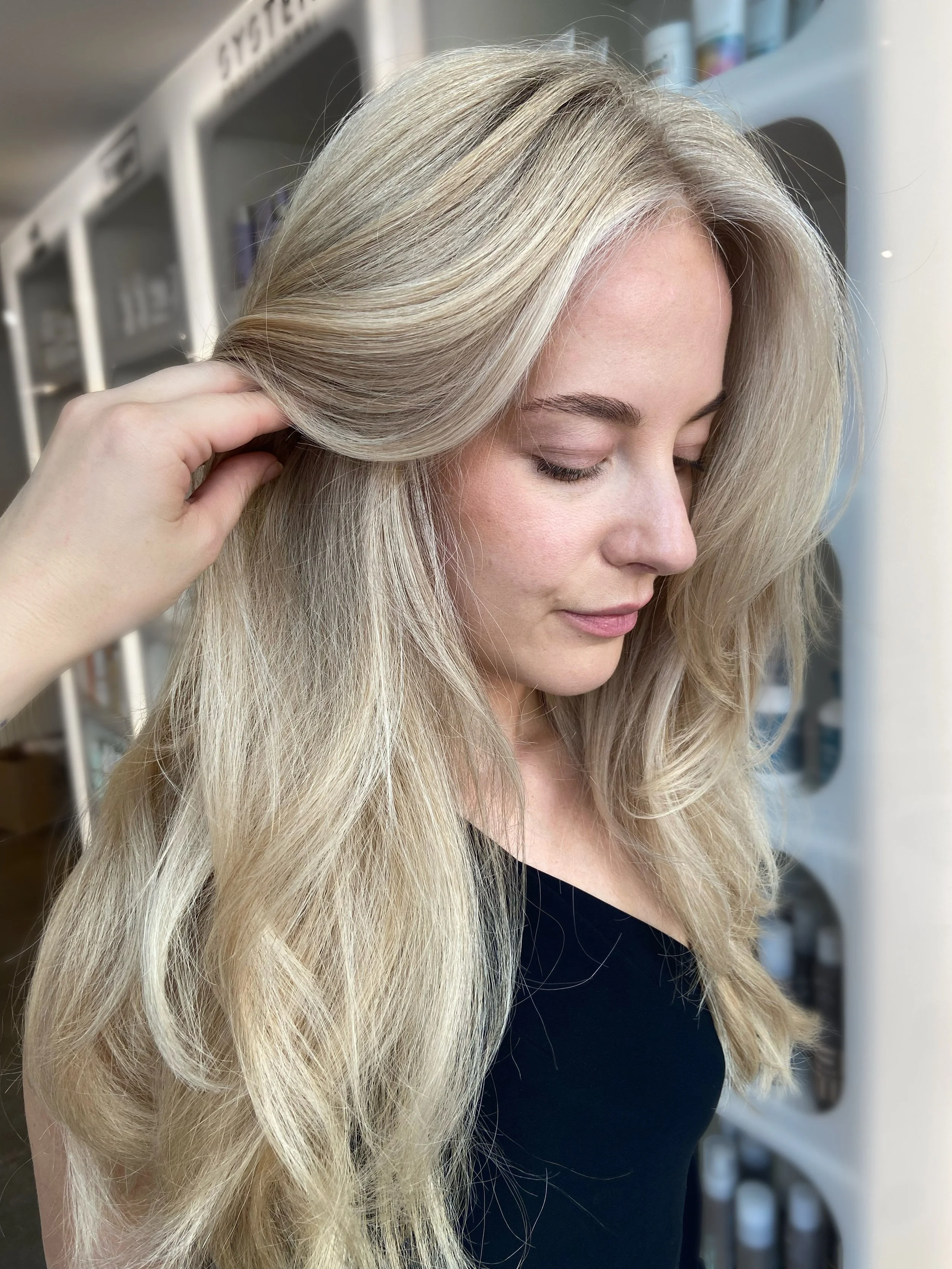 Close-up of a woman with long, blonde hair, gently touching her hair with her hand, in a salon setting.