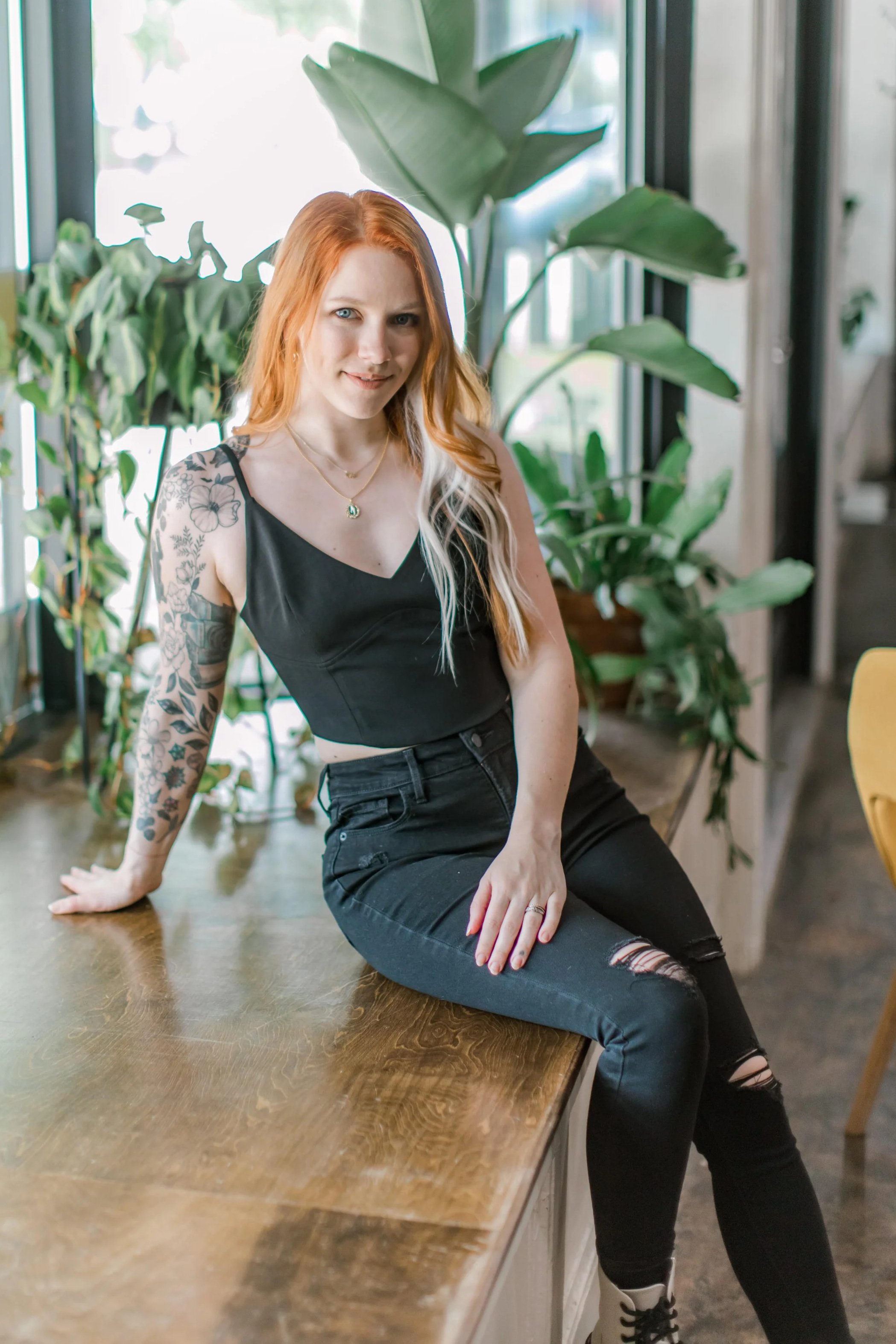 A young woman with red hair and tattoos on her arm, sitting on a wooden table near large green plants, wearing a black sleeveless top and ripped black jeans, in a brightly lit room with windows. alternative hairstylist