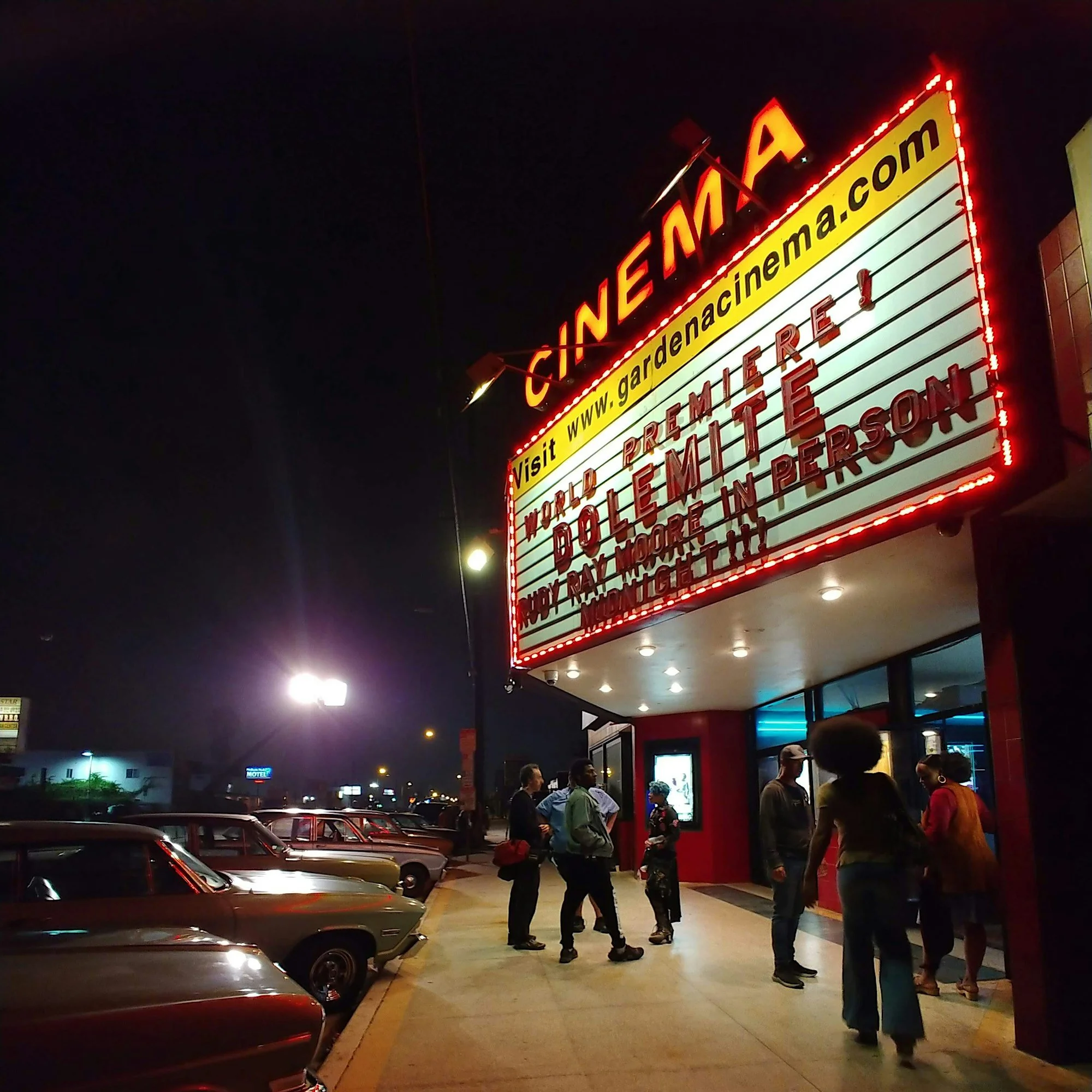 People standing outside the Gardena Cinema at night, with cars parked nearby and the digital marquee displaying movie titles. It's the premiere of Dolemite is My Name.